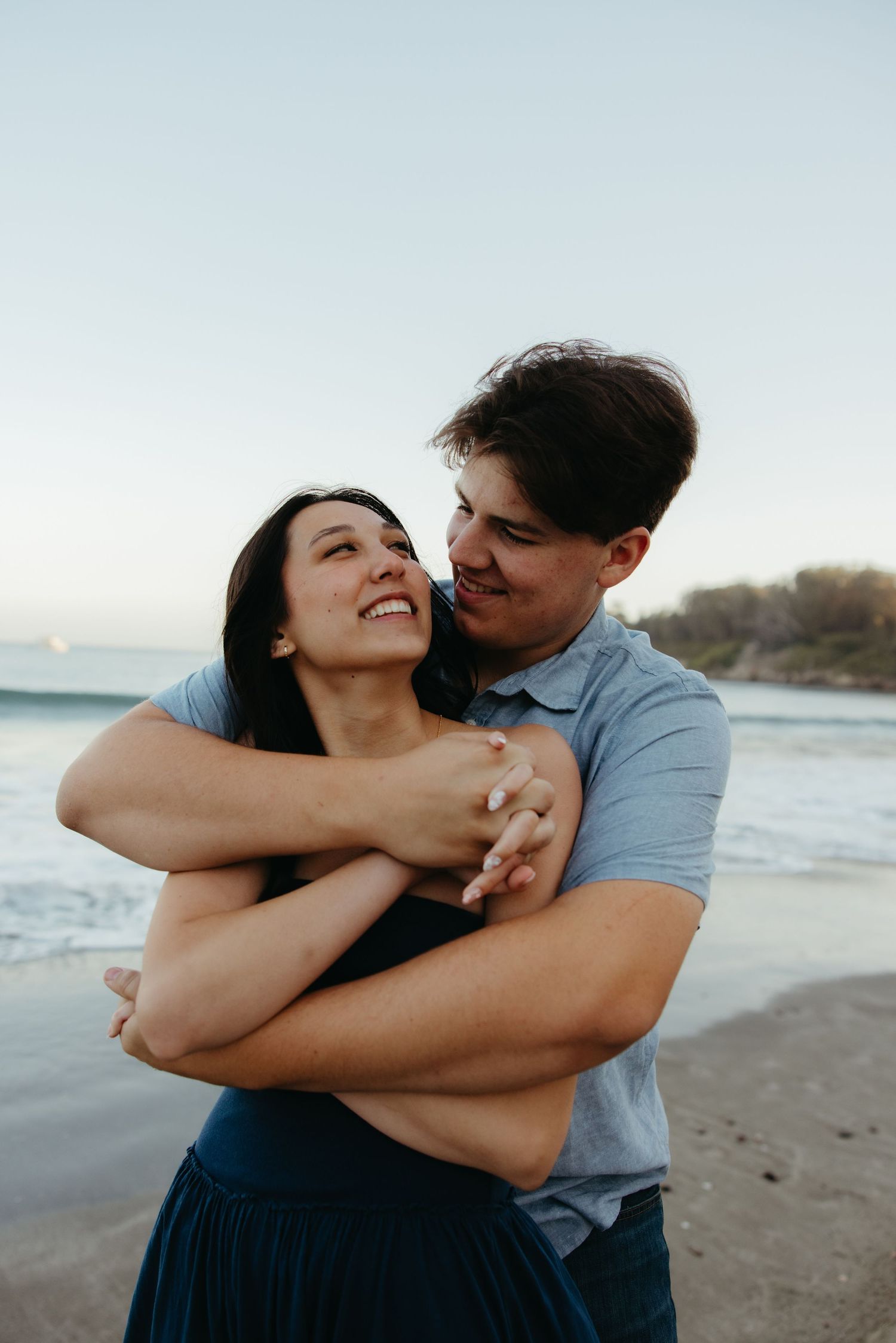 Happy couple embraces and laughs together on a sandy beach at dusk with ocean waves behind them.