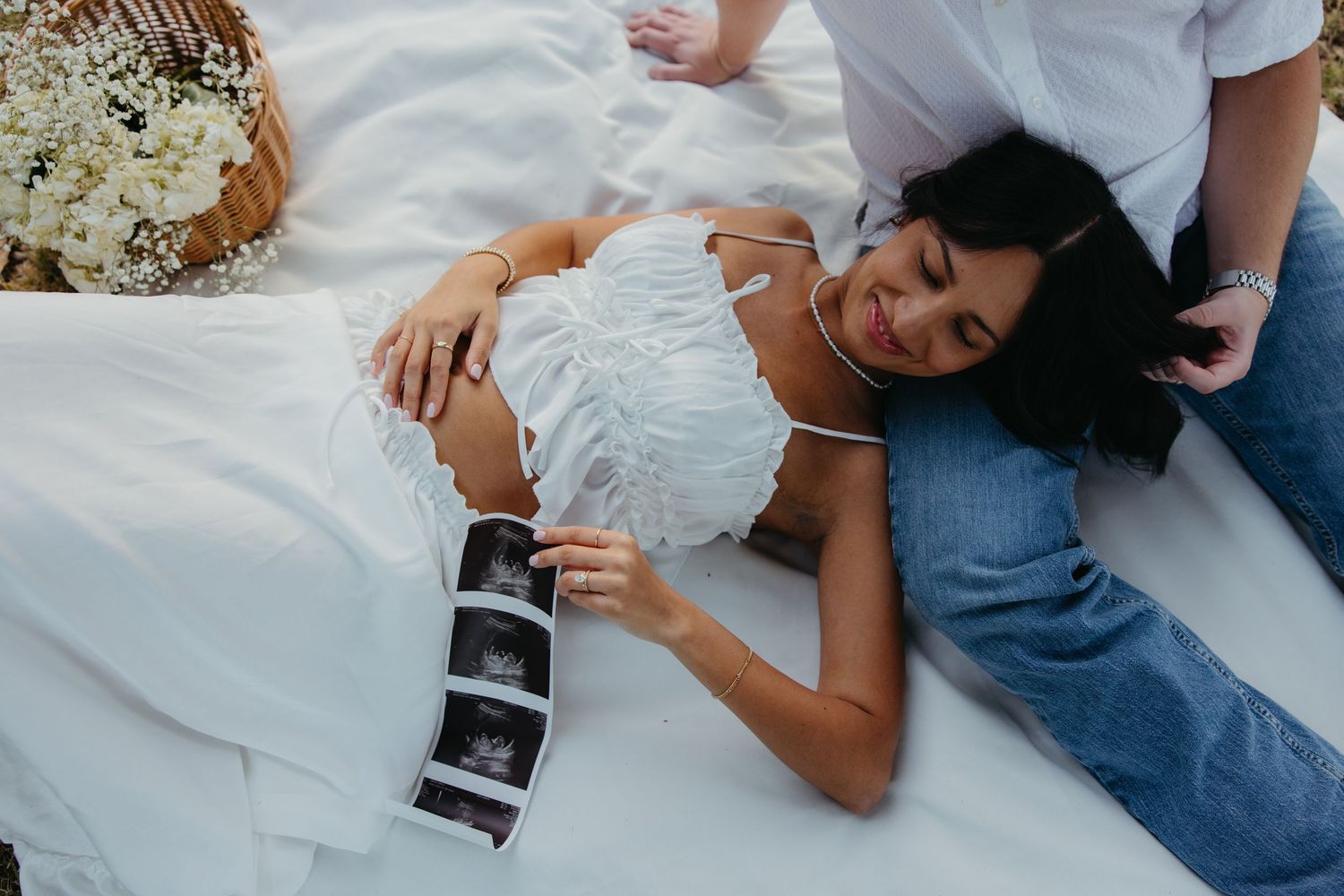 A pregnant woman lies on a white blanket holding ultrasound photos while her partner sits beside her.