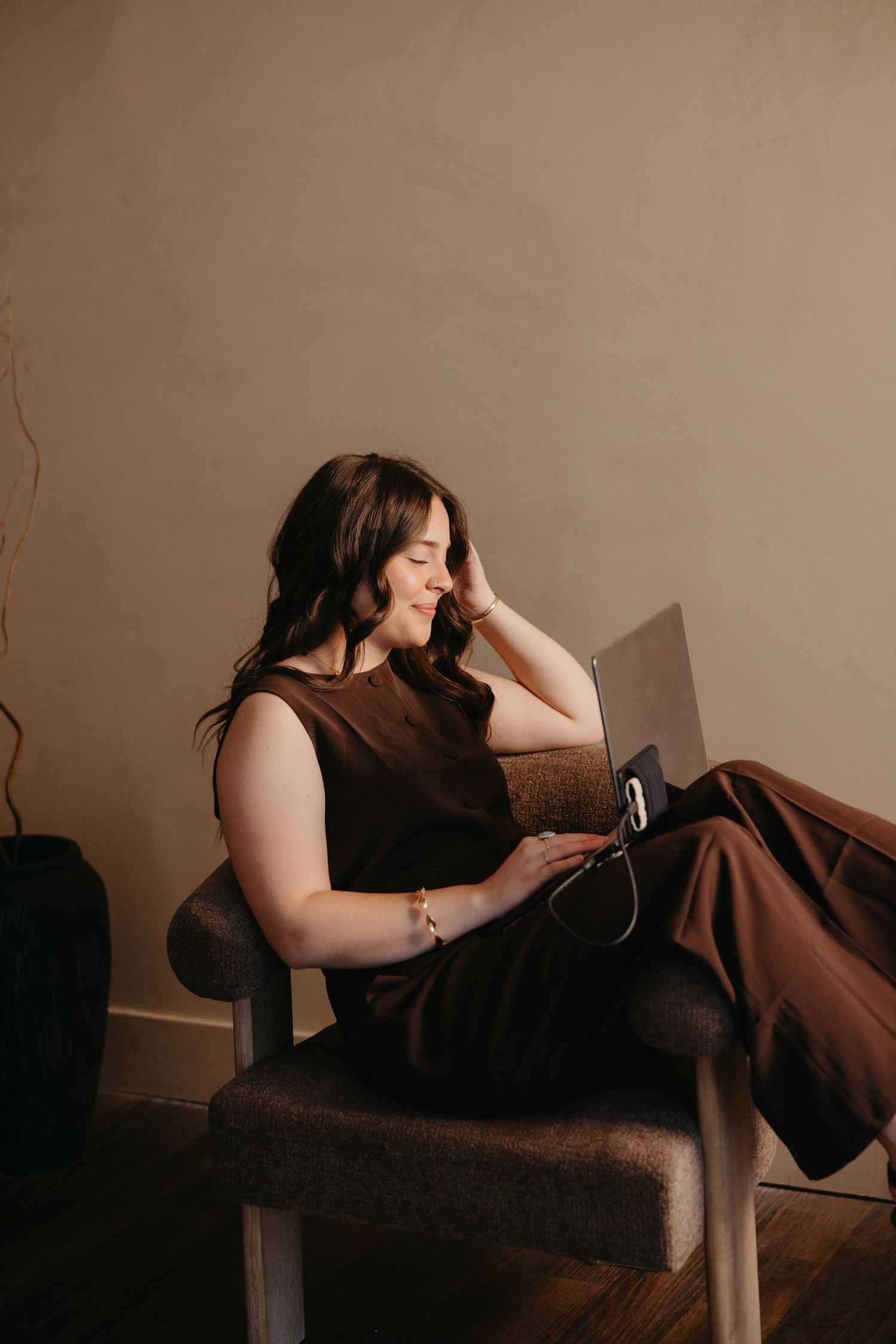 Woman in brown outfit smiling while working on a laptop in a modern chair.