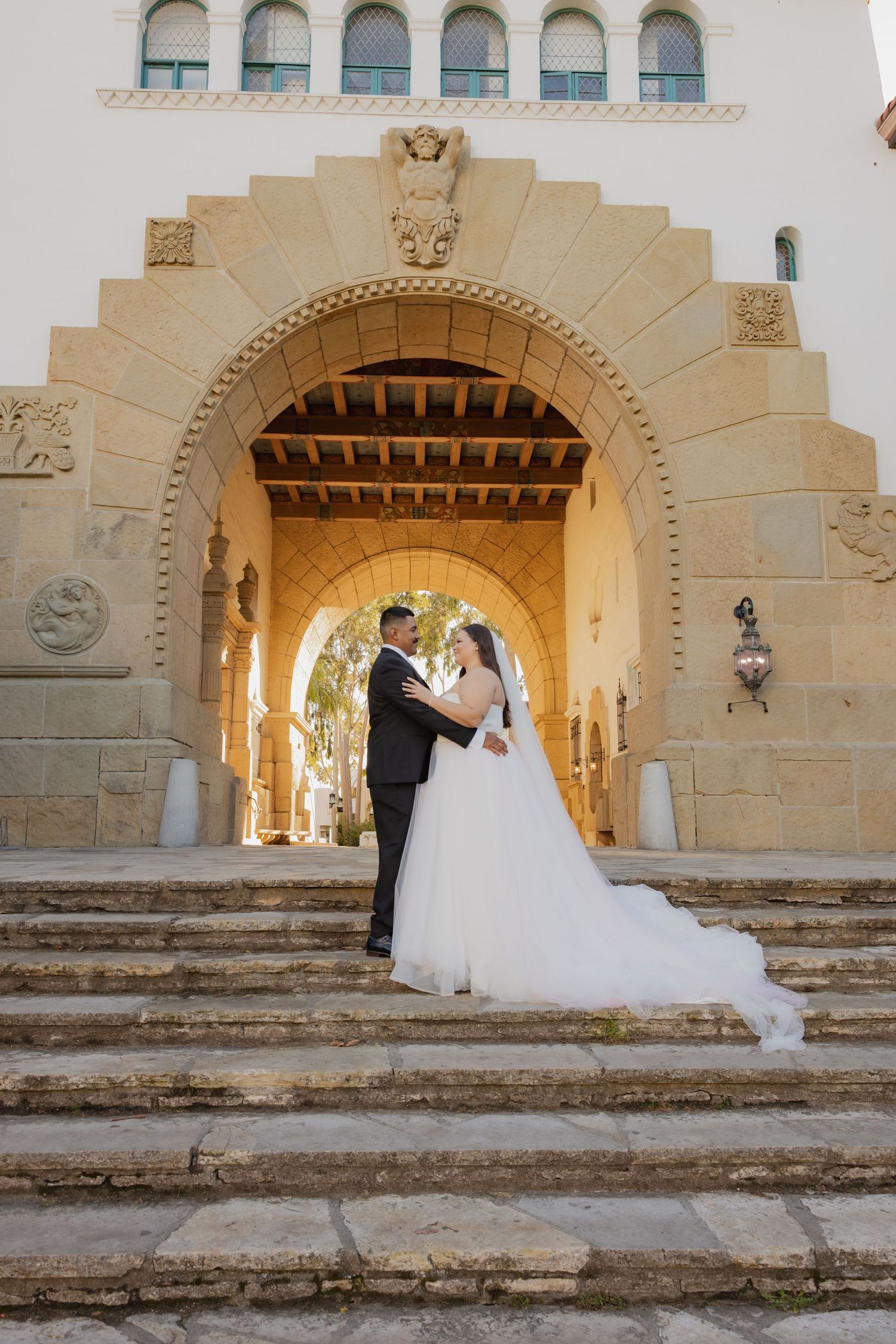 Bride and groom embracing under an ornate stone archway on steps of a historic building.