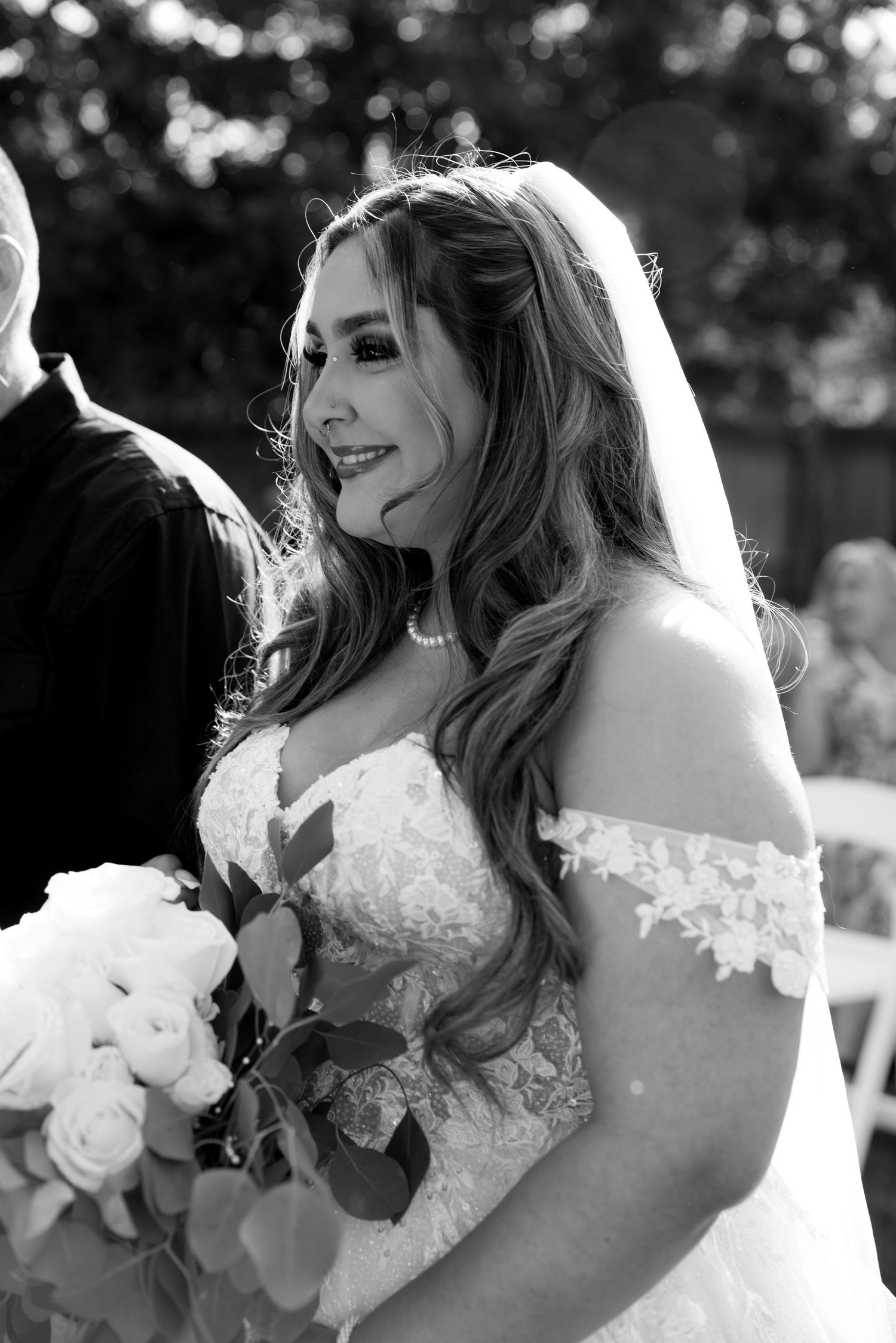 Radiant bride smiling in off-shoulder lace wedding gown and veil during outdoor wedding ceremony.