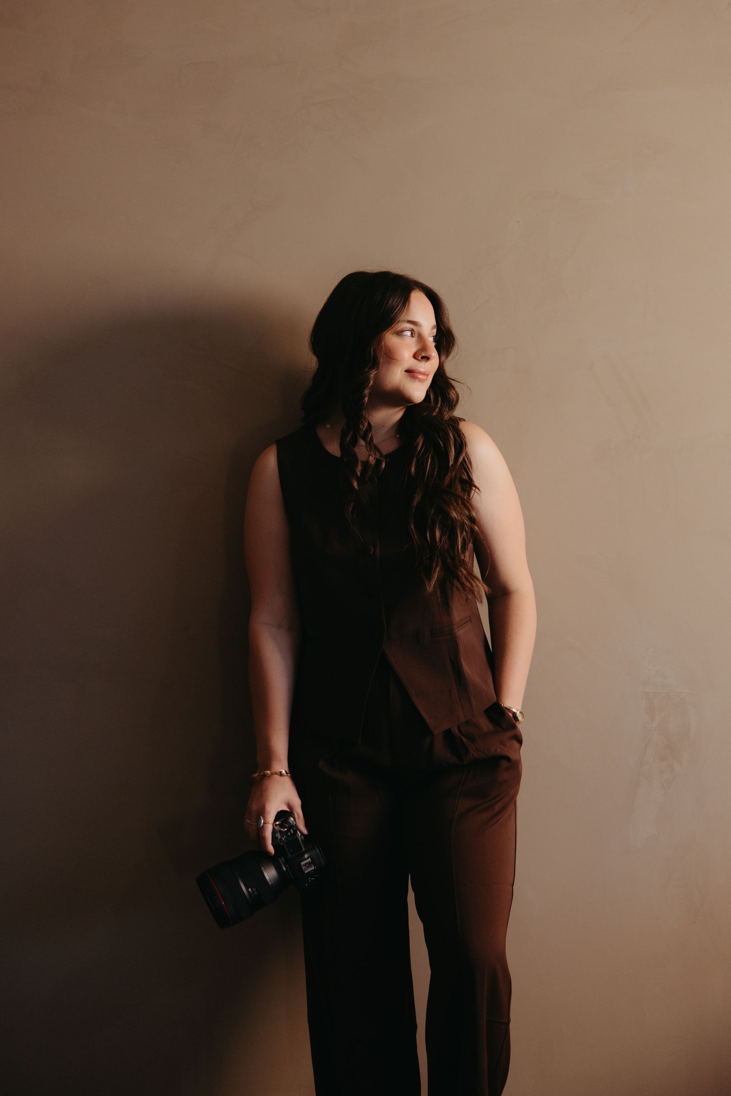 Brunette woman in black sleeveless top holding a DSLR camera while leaning against a beige wall.