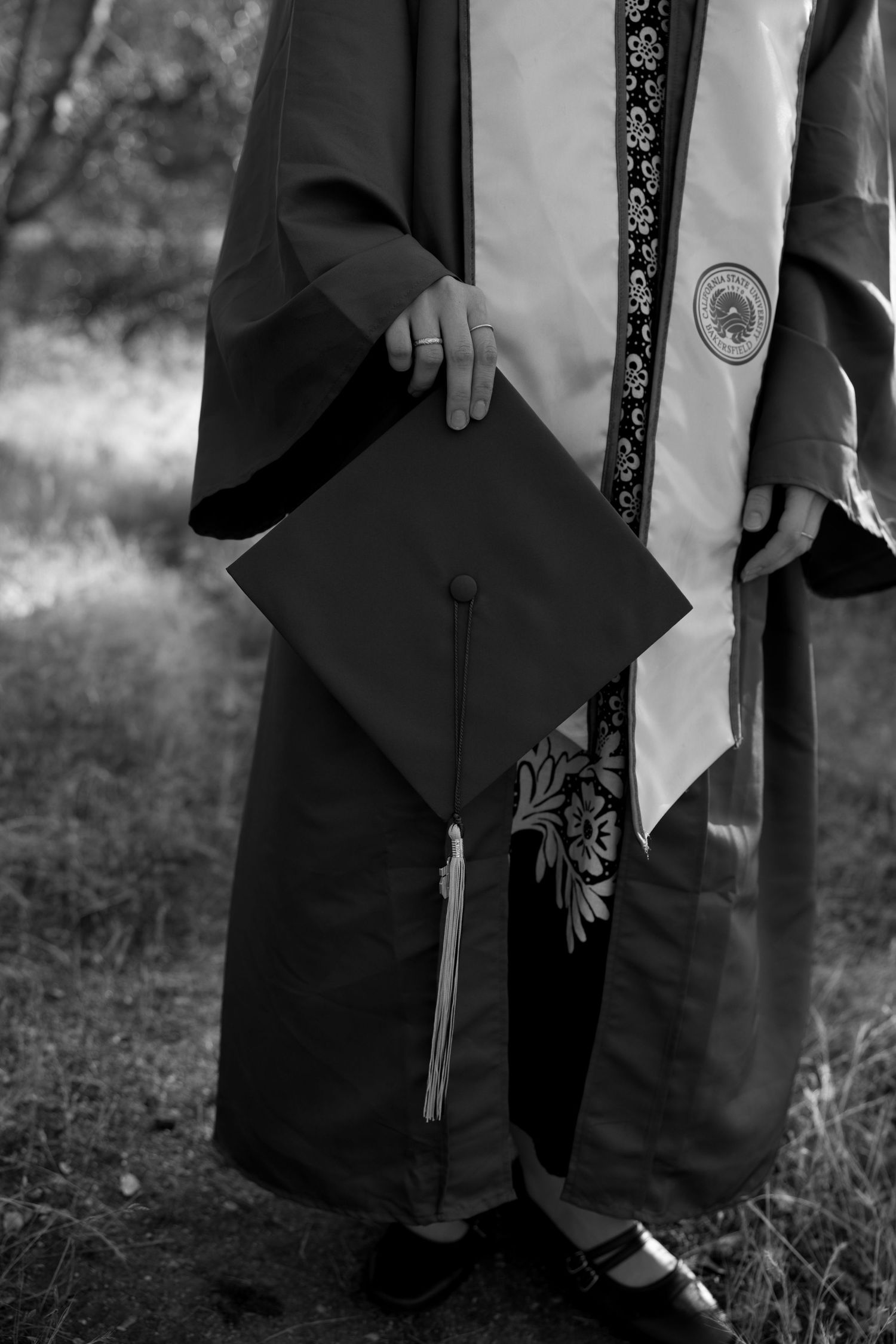 Black and white close-up of graduate in cap and gown holding mortarboard outdoors.