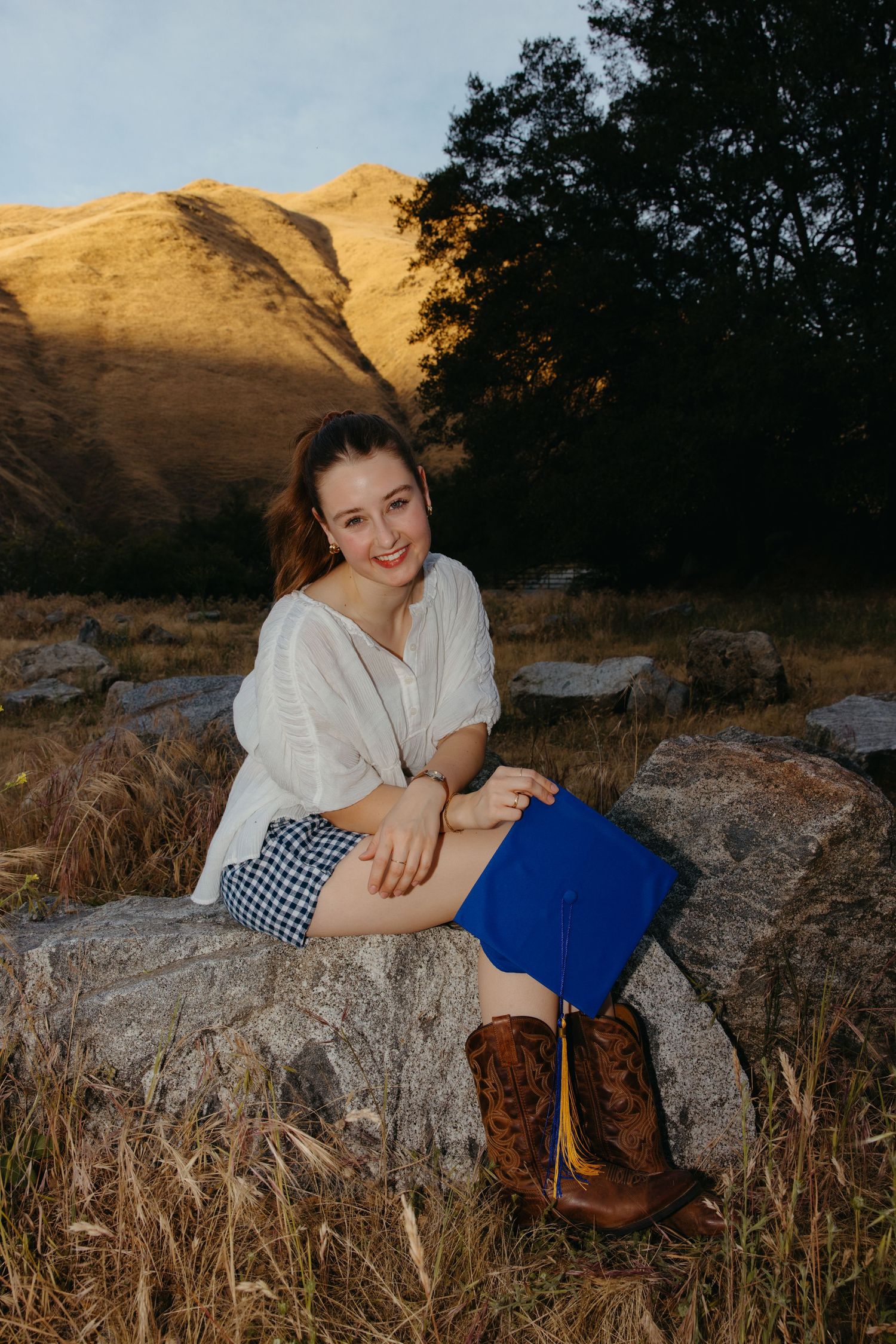 Smiling graduate in white top and shorts holding blue cap while sitting on rocks with mountain backdrop.