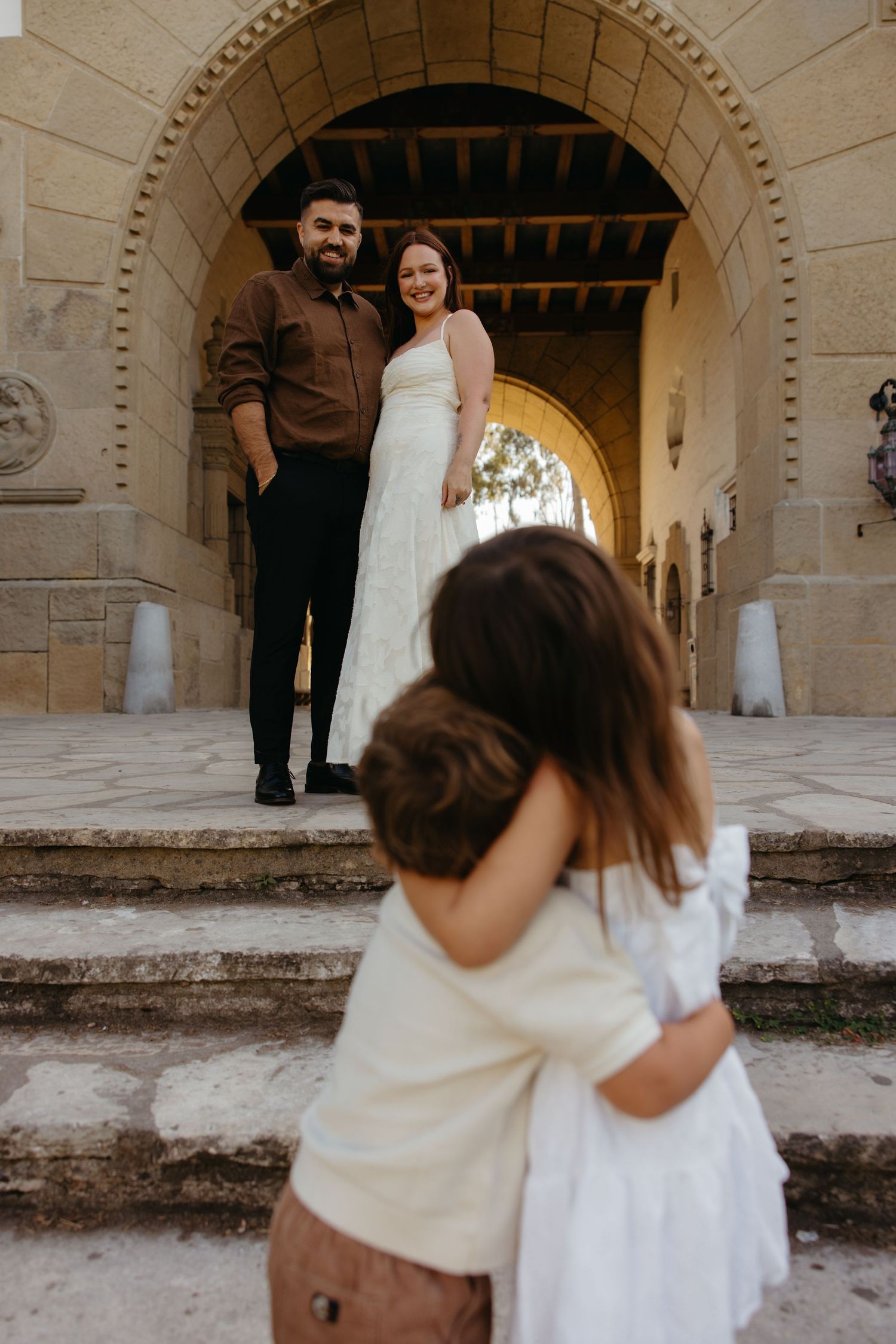 Couple in wedding attire smiling on steps as two children run toward them under stone arch.
