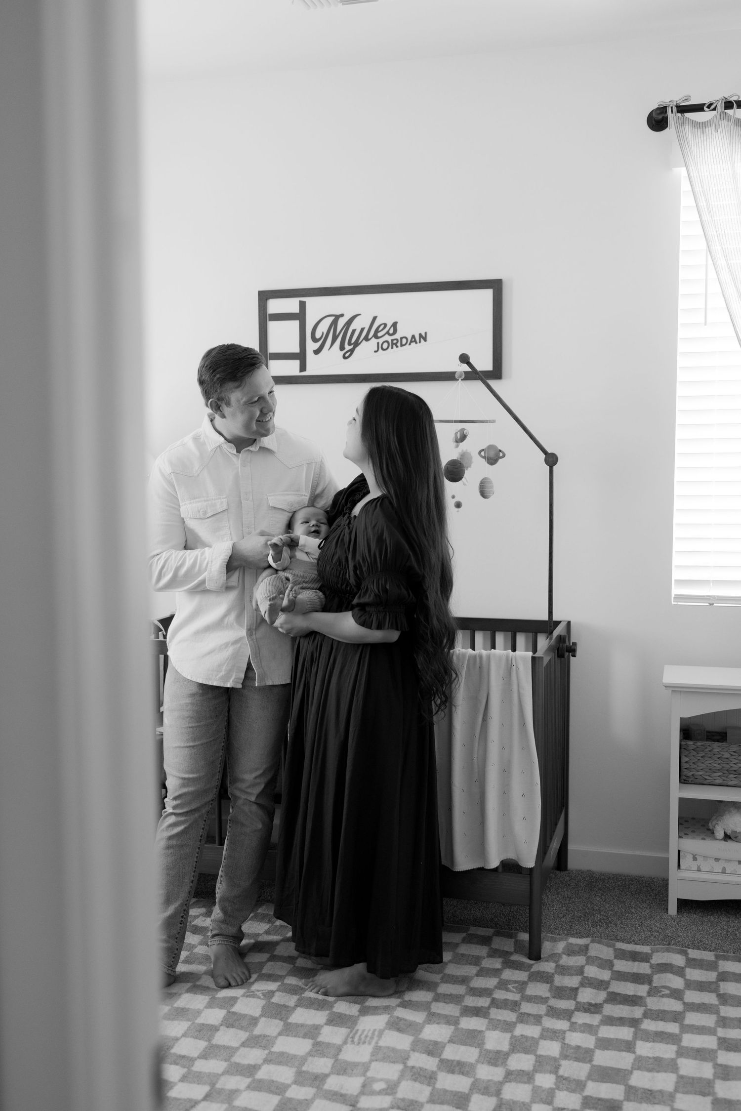 Black and white photo of new parents holding their newborn in a nursery with a Myles Jordan sign.