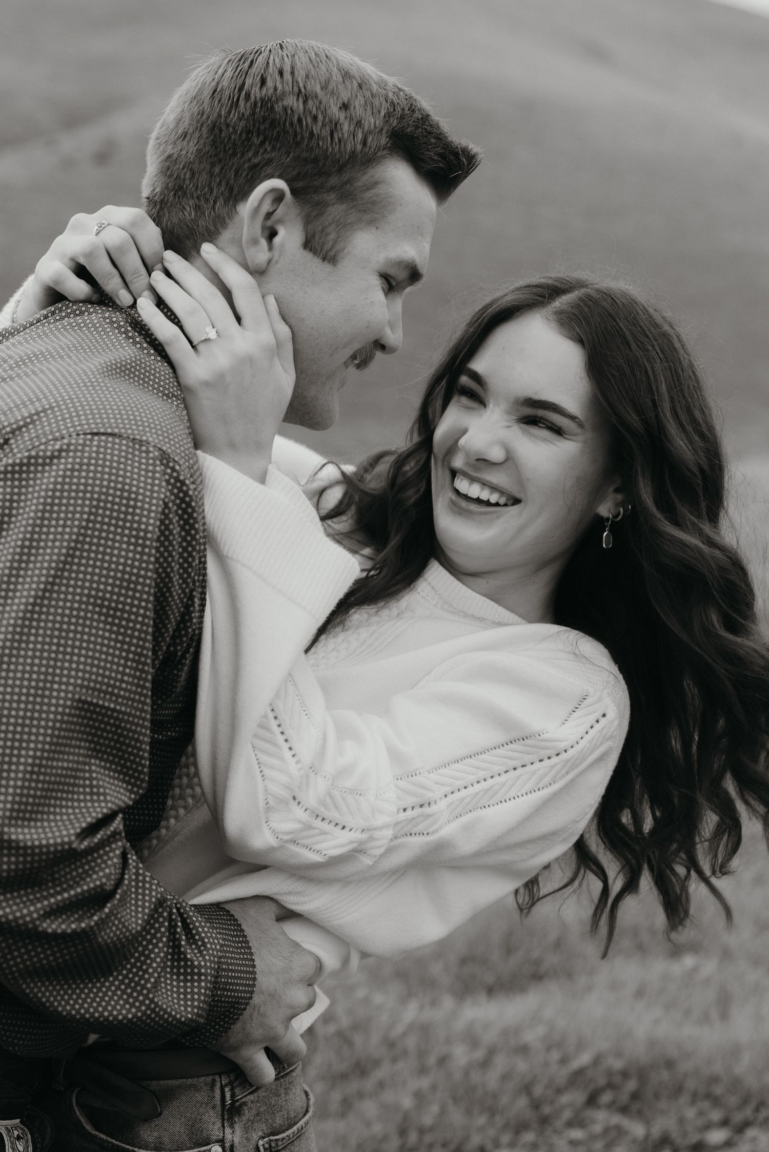 Black and white photo of a smiling woman with wavy hair embracing a man in a grassy field.