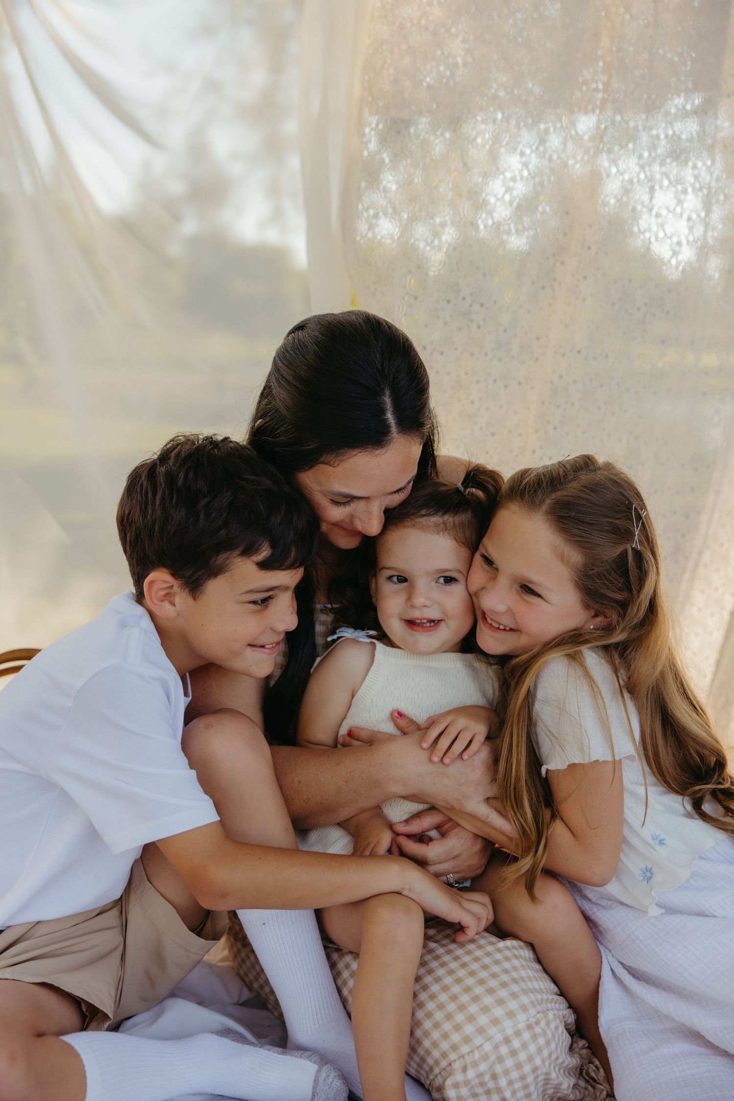 A mother hugs her three happy children together under soft draped fabric in warm light.