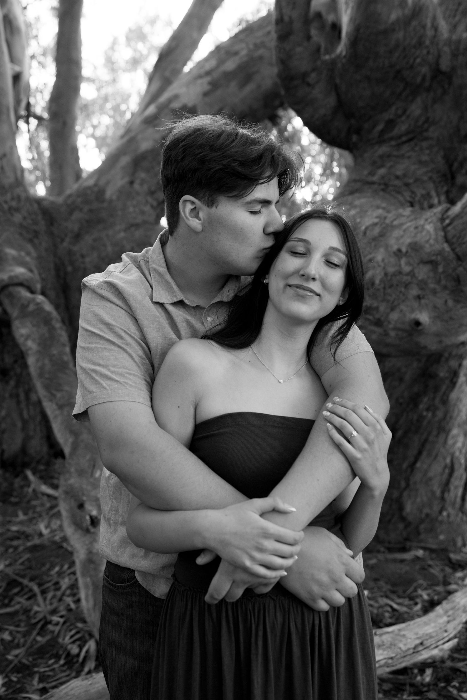 Black and white photo of couple embracing tenderly in front of a large tree, man kissing womans temple.