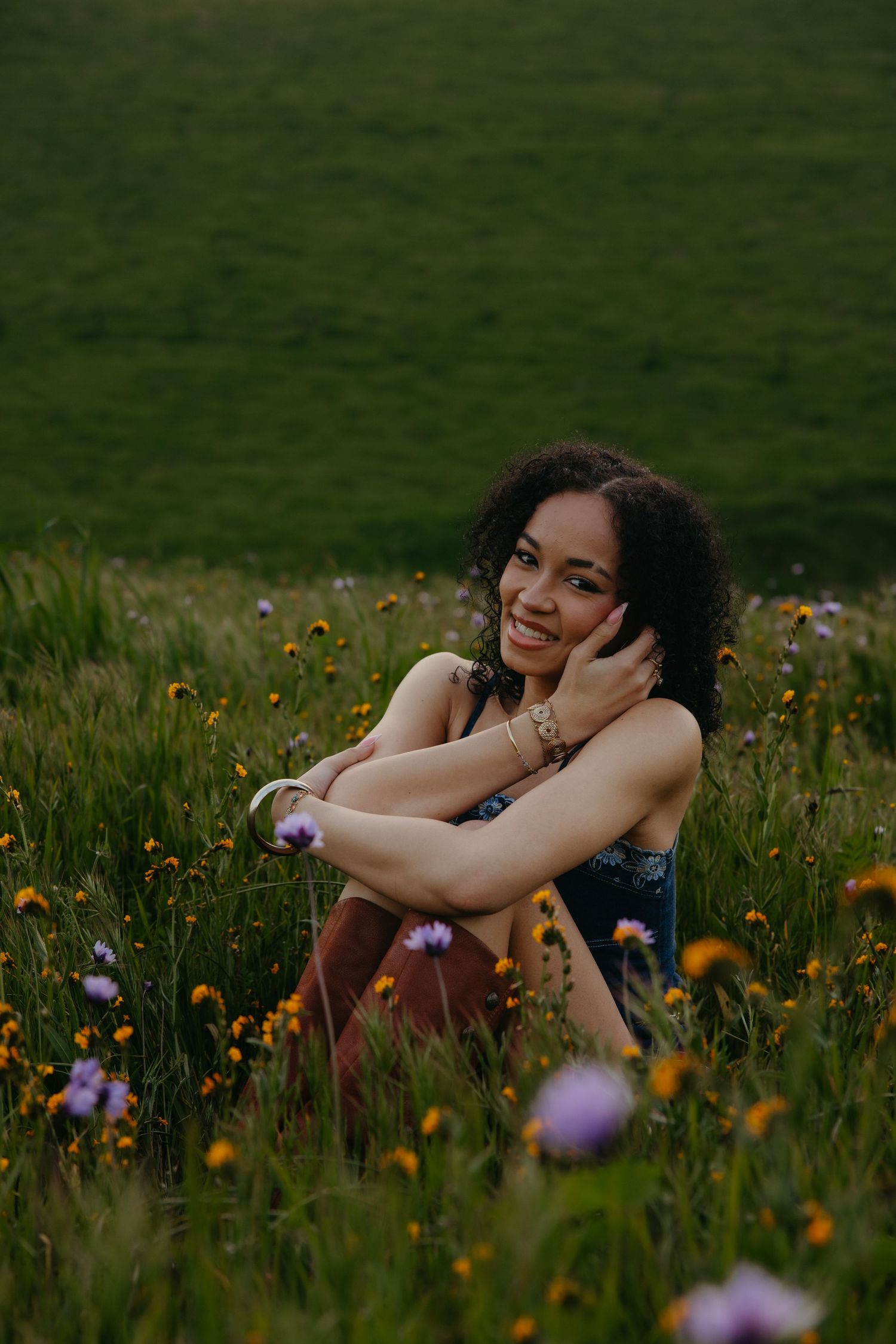 Smiling young woman with curly hair sits relaxed in a colorful wildflower field at dusk, hugging her knees.