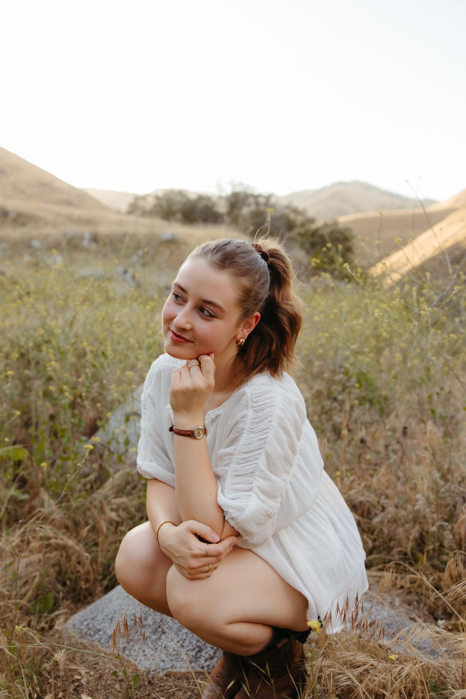 Young woman in white top crouching on rock smiling in golden sunlit hillside meadow.