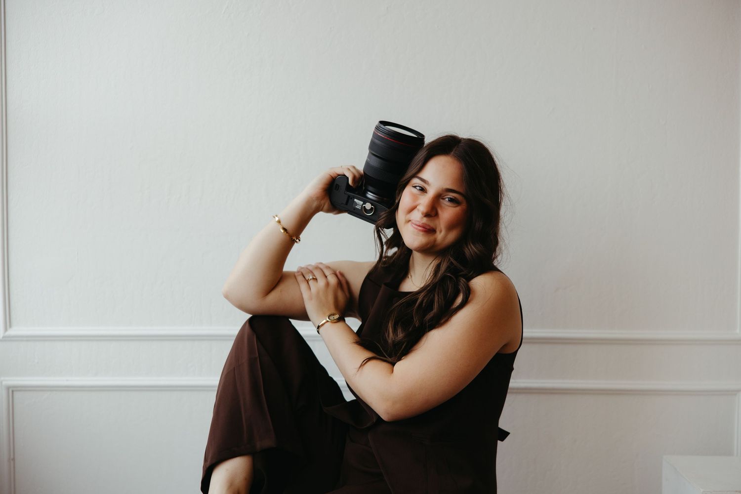 Young woman sitting against white wall holding a camera lens, smiling, wearing dark outfit, photographer portrait.