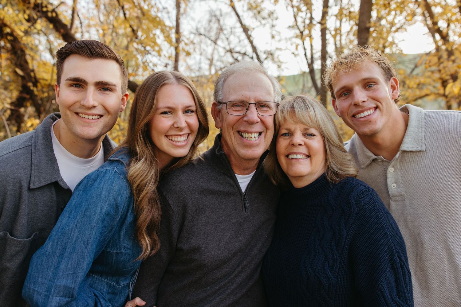 Smiling family of five posing together outdoors surrounded by vibrant autumn foliage.