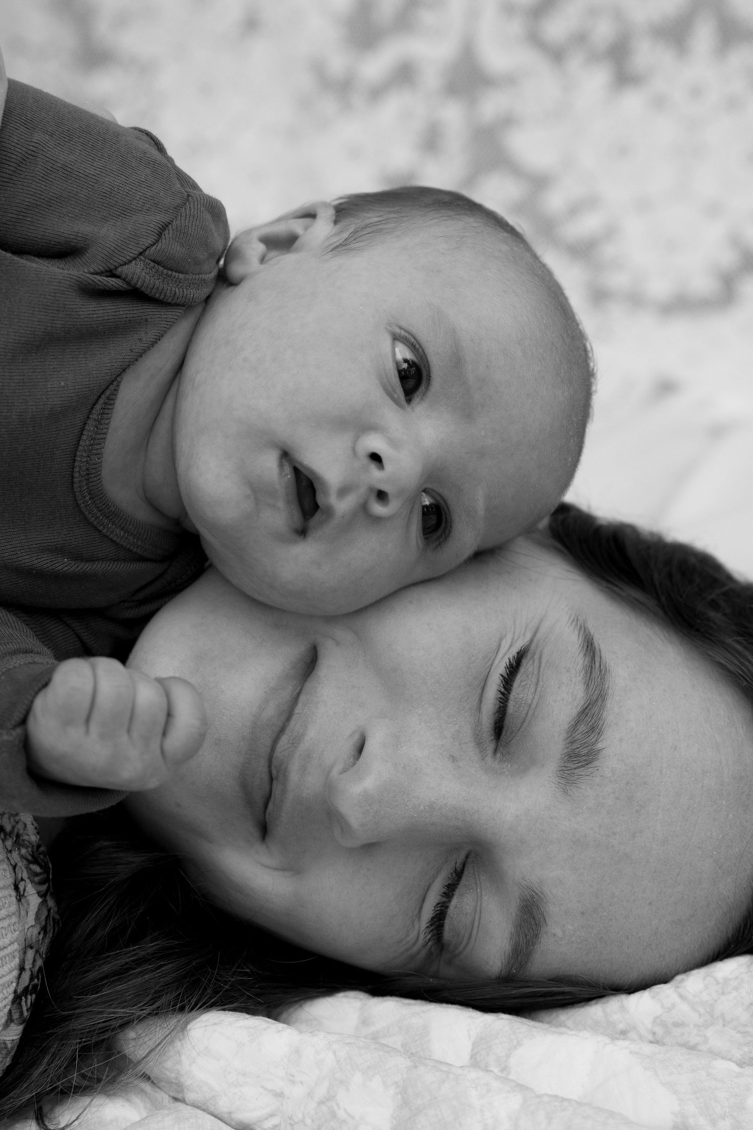 Black and white photo of a sleeping mother with her curious baby resting on her head, sharing a tender moment.