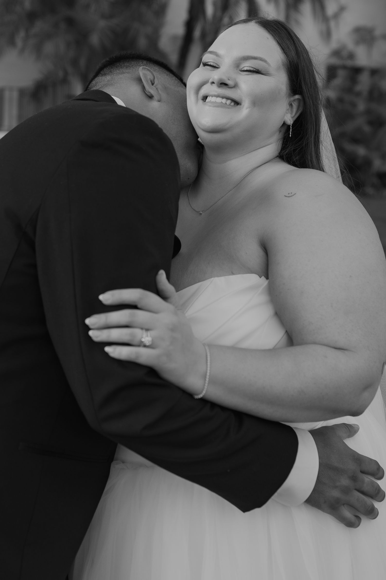 Black and white photo of a smiling bride embracing her groom during outdoor wedding portraits.