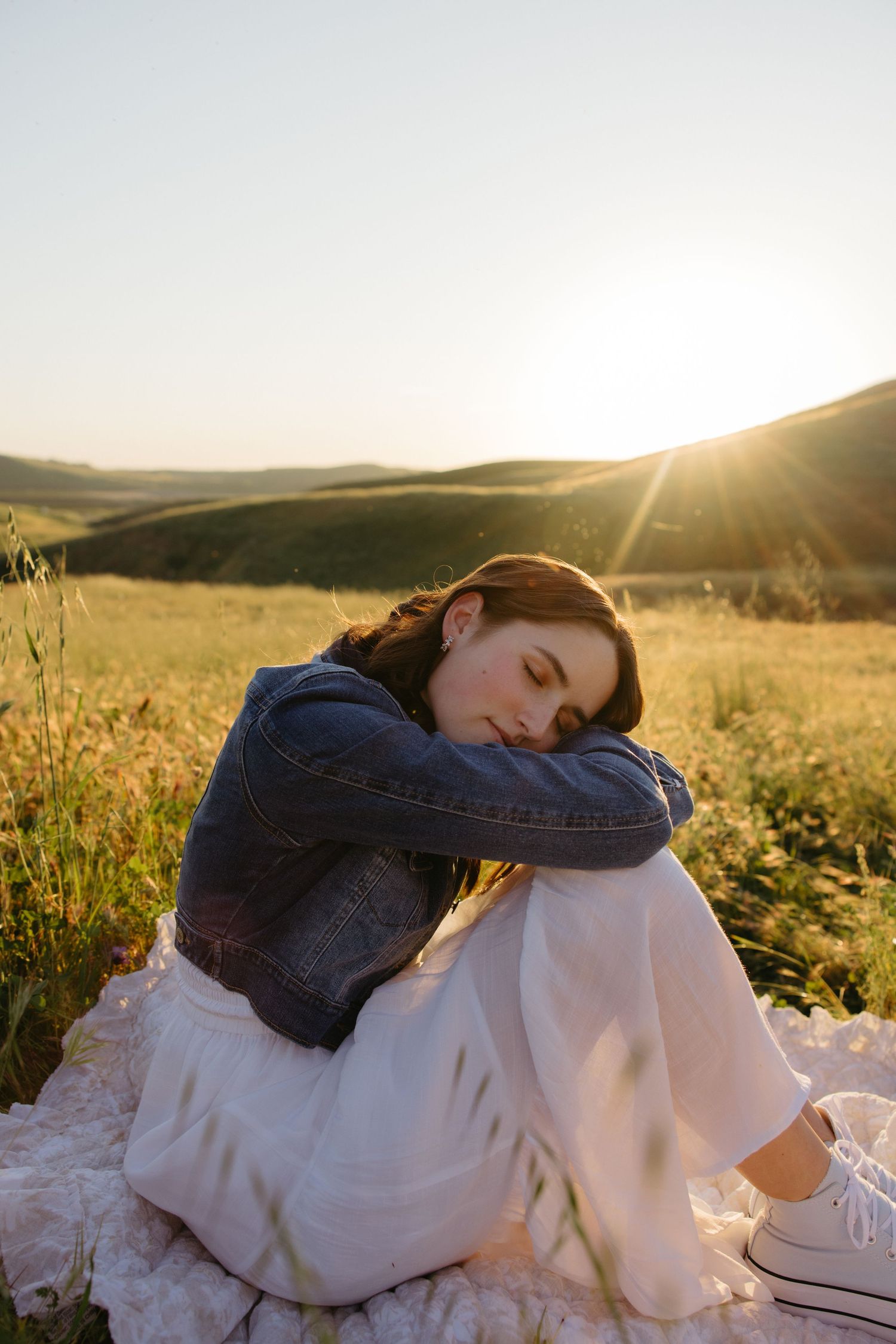 Young woman in denim jacket sitting peacefully in a sunlit golden meadow at sunset.