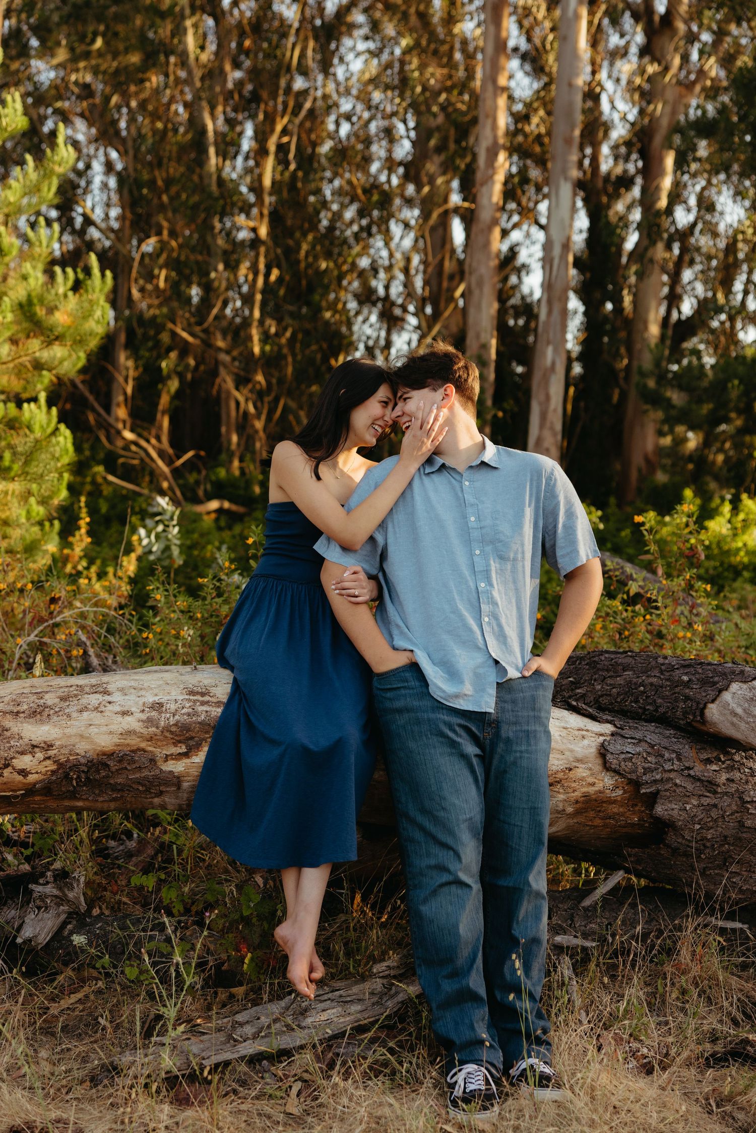 Couple sharing a close romantic moment leaning against a fallen log surrounded by tall trees in warm sunlight.