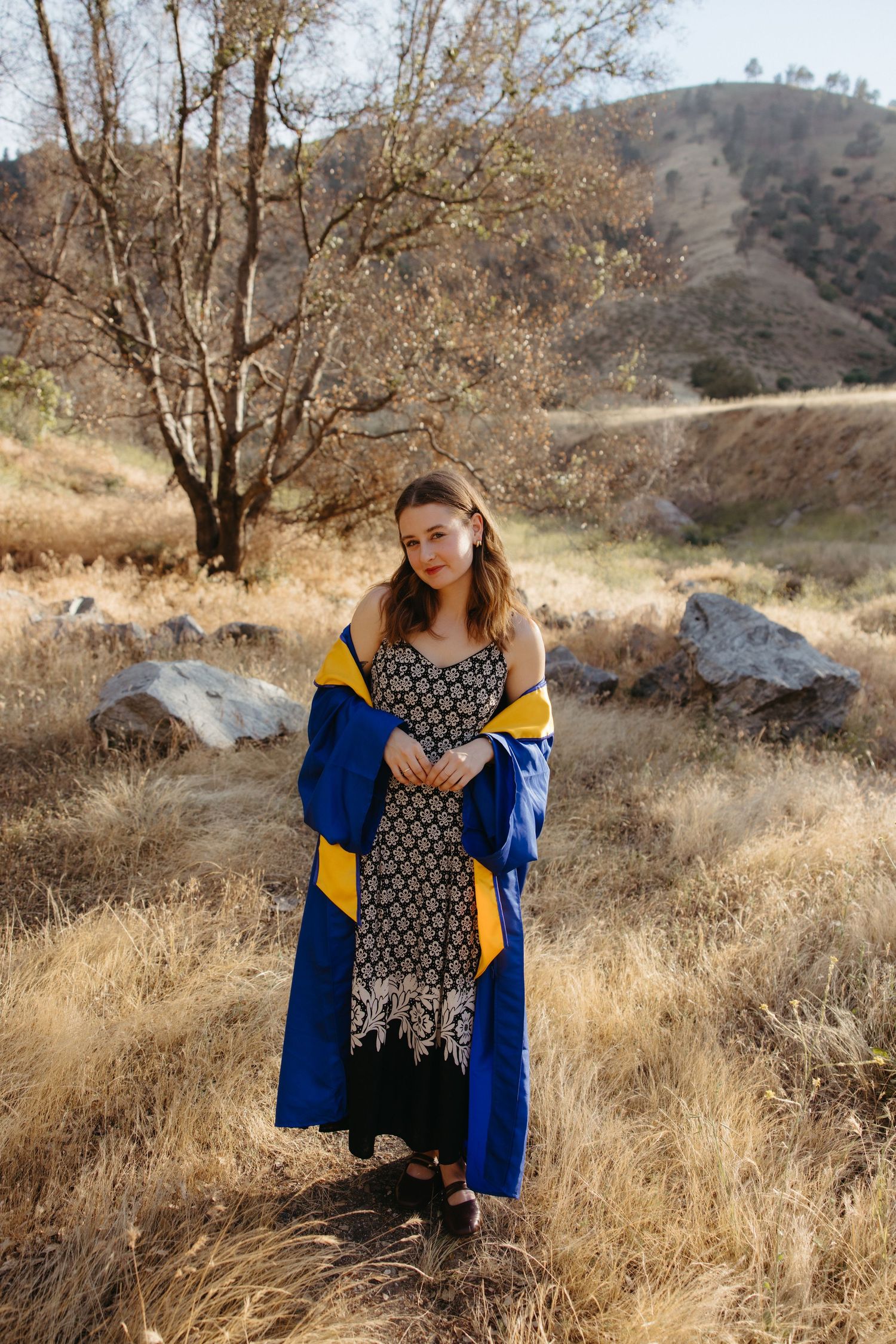Woman in black floral dress wearing blue and gold graduation regalia outdoors in dry grassy field.