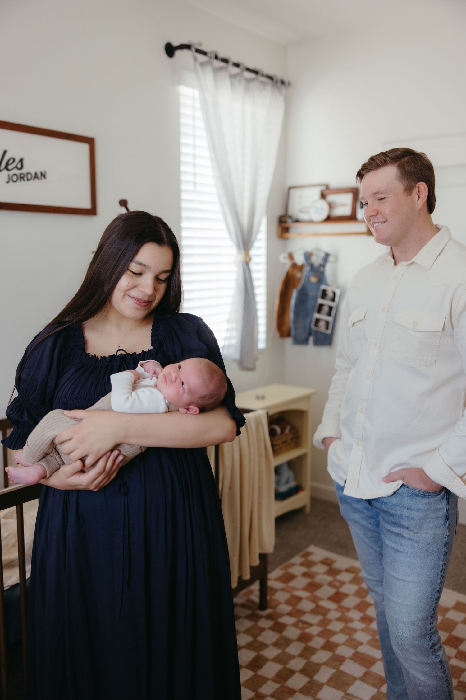 New mother holds newborn baby while smiling father stands beside her in a cozy nursery.