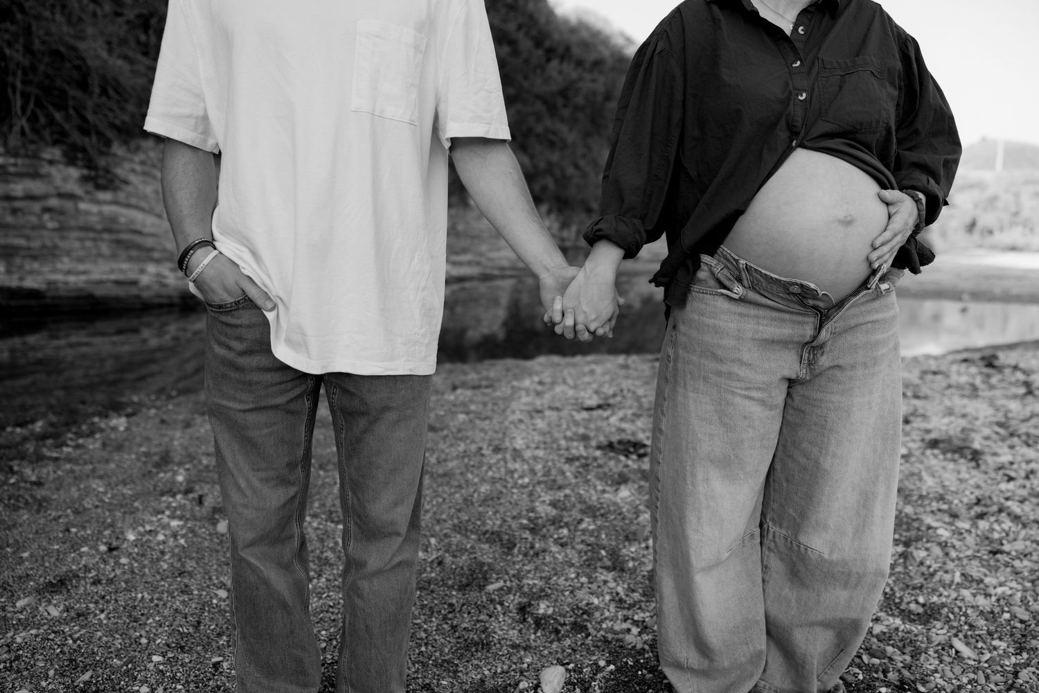 Black and white photo of pregnant couple holding hands on a rocky beach, bump visible.