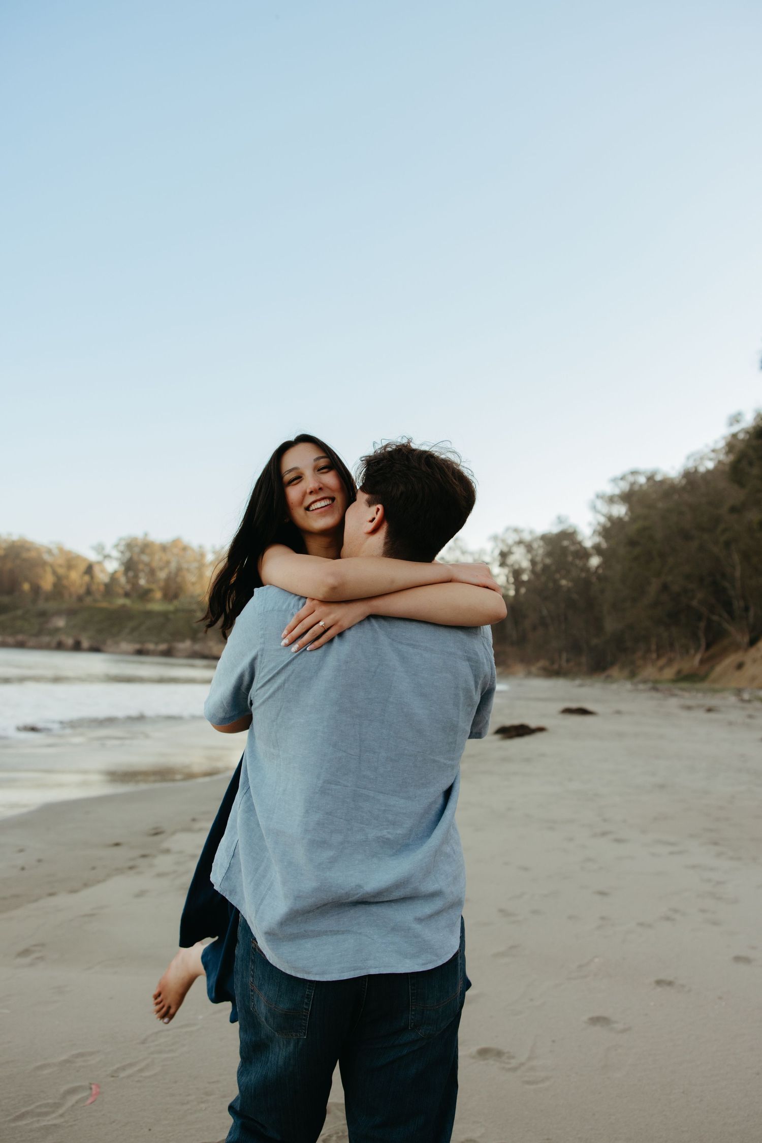 Happy couple embracing and laughing on a sandy beach at sunset with trees in background.