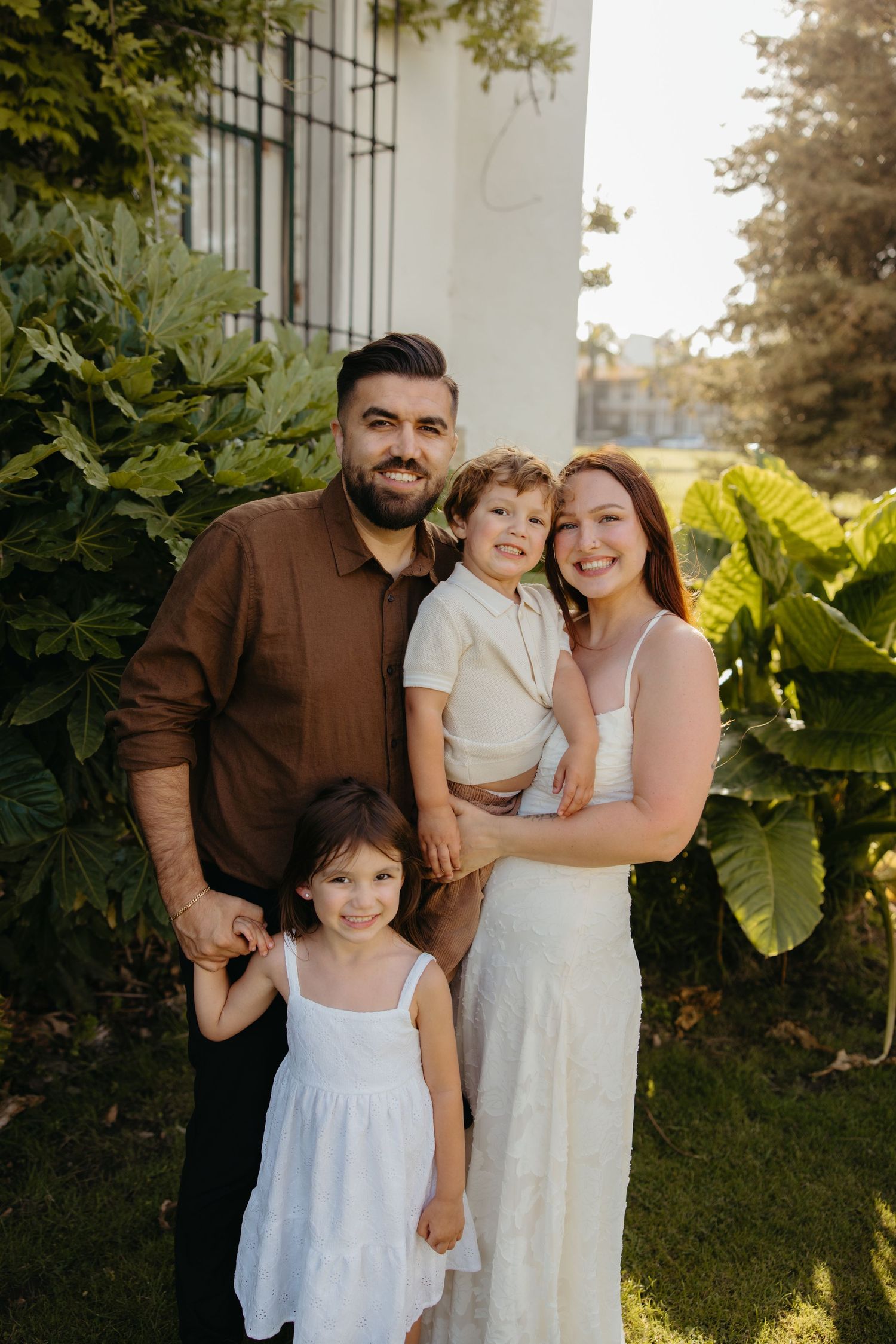 Smiling family of four posing together in golden sunlight beside tropical green plants.