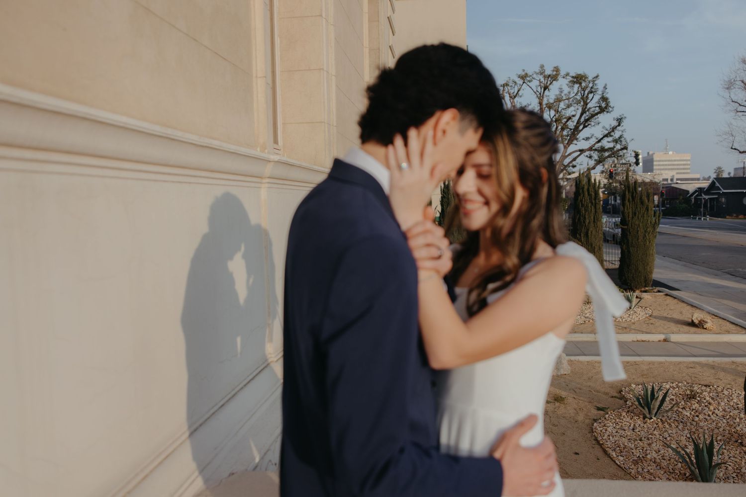 A smiling bride in a white dress embraces a groom in a navy suit against a sunlit building wall.