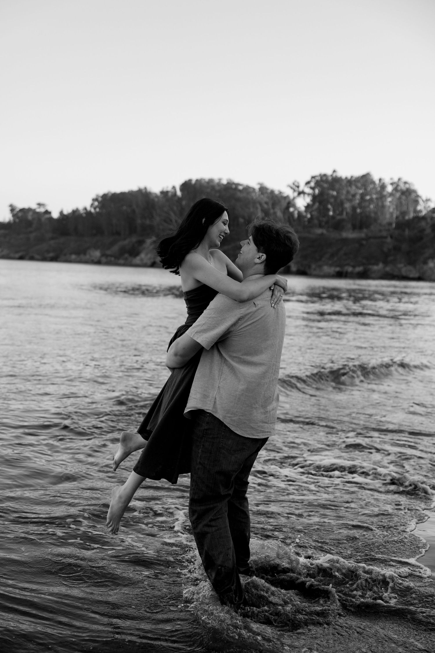 Black and white photo of a man lifting a laughing woman in his arms while standing in a river.