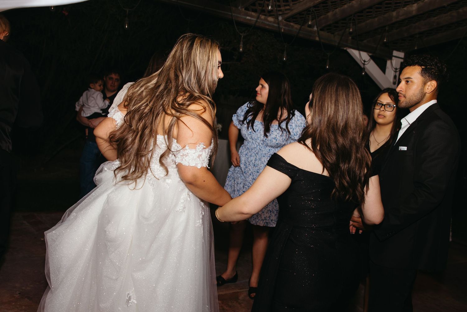 Bride in off-shoulder gown dances joyfully with guests at a lively outdoor wedding reception.