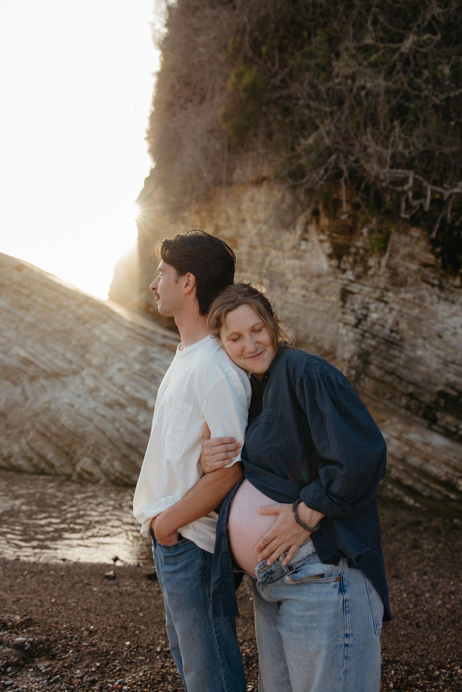 Pregnant woman leans against partner while cradling baby bump on scenic rocky beach at sunset.