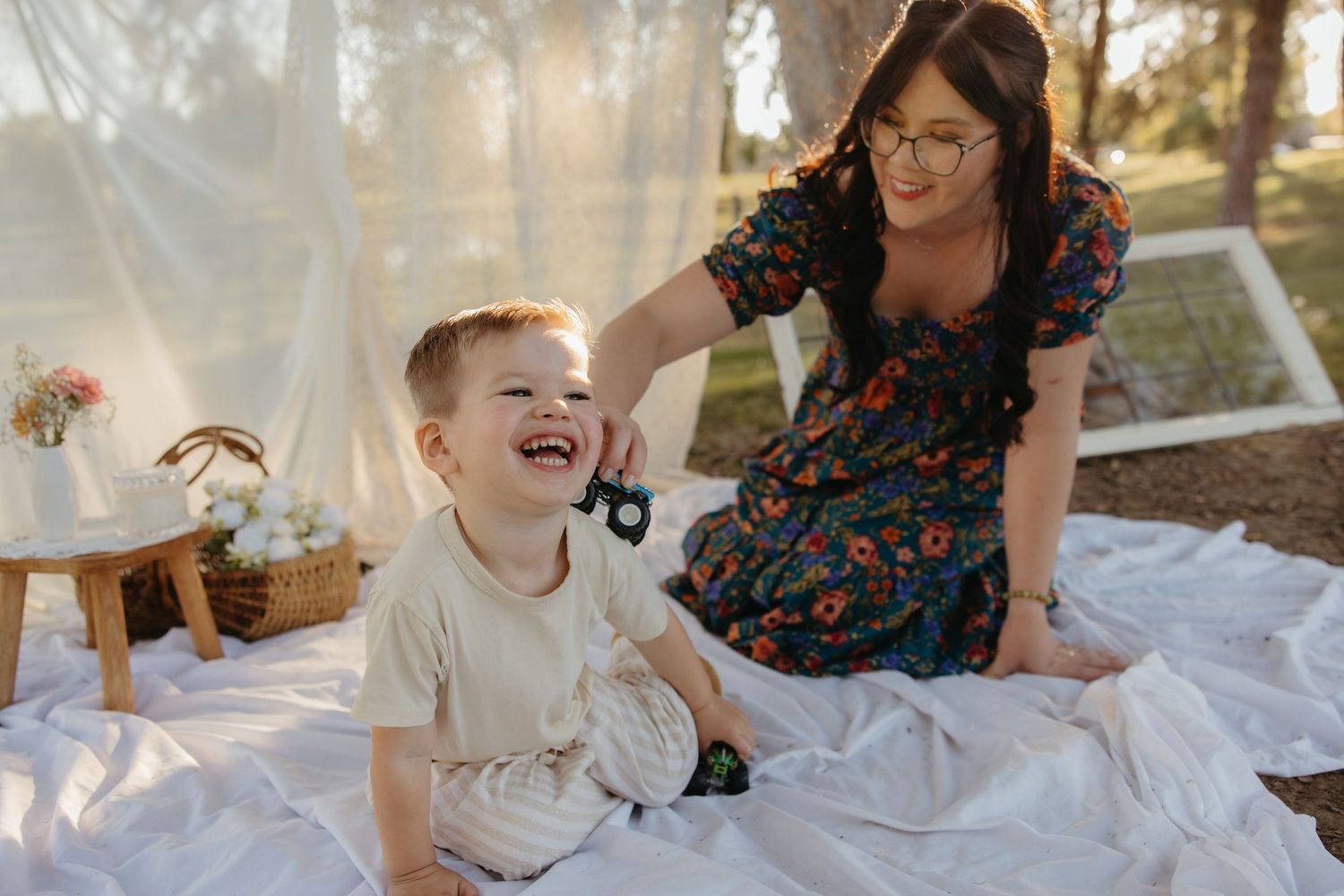 A laughing young boy sits on a white blanket outdoors while his mother smiles and plays with him.