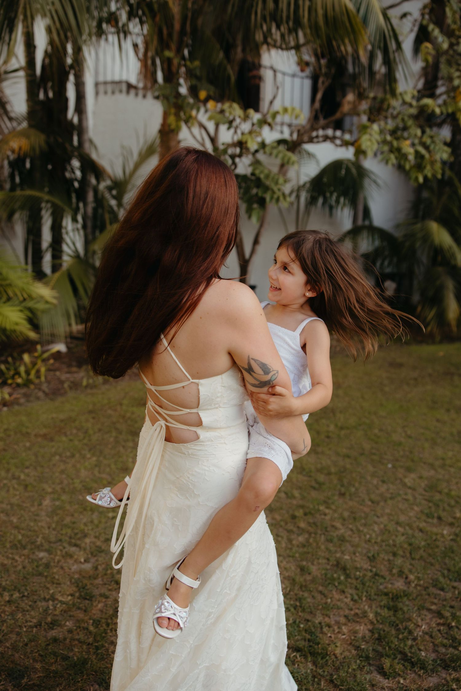 Mother in white wedding gown holding young daughter while spinning outdoors near palm trees.