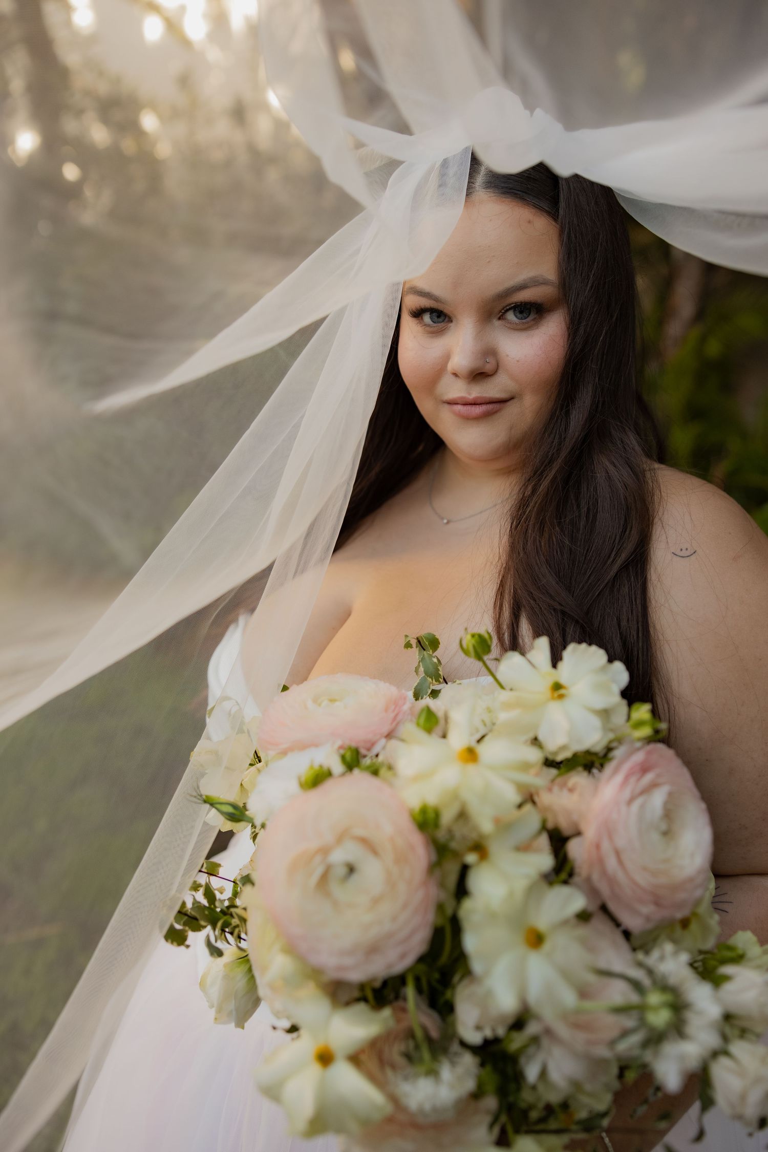 Bride holding a pink and white floral bouquet under a flowing veil in soft golden light.
