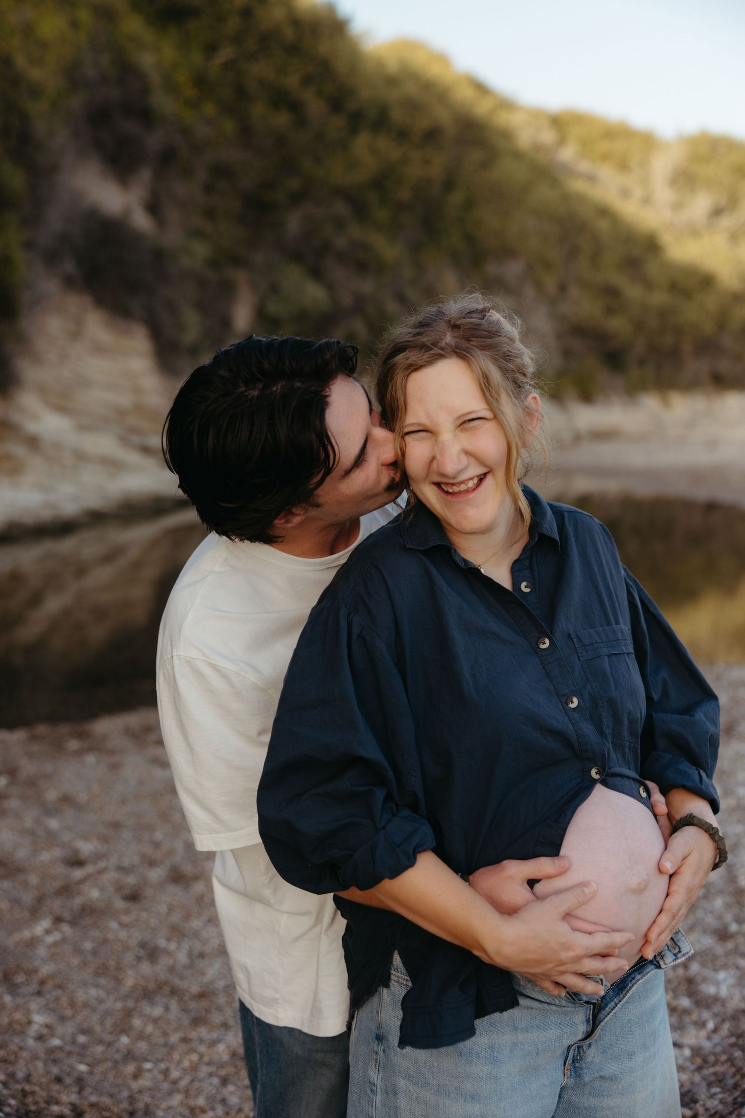 Man kisses pregnant woman's cheek as she laughs joyfully on a pebble beach at golden hour.