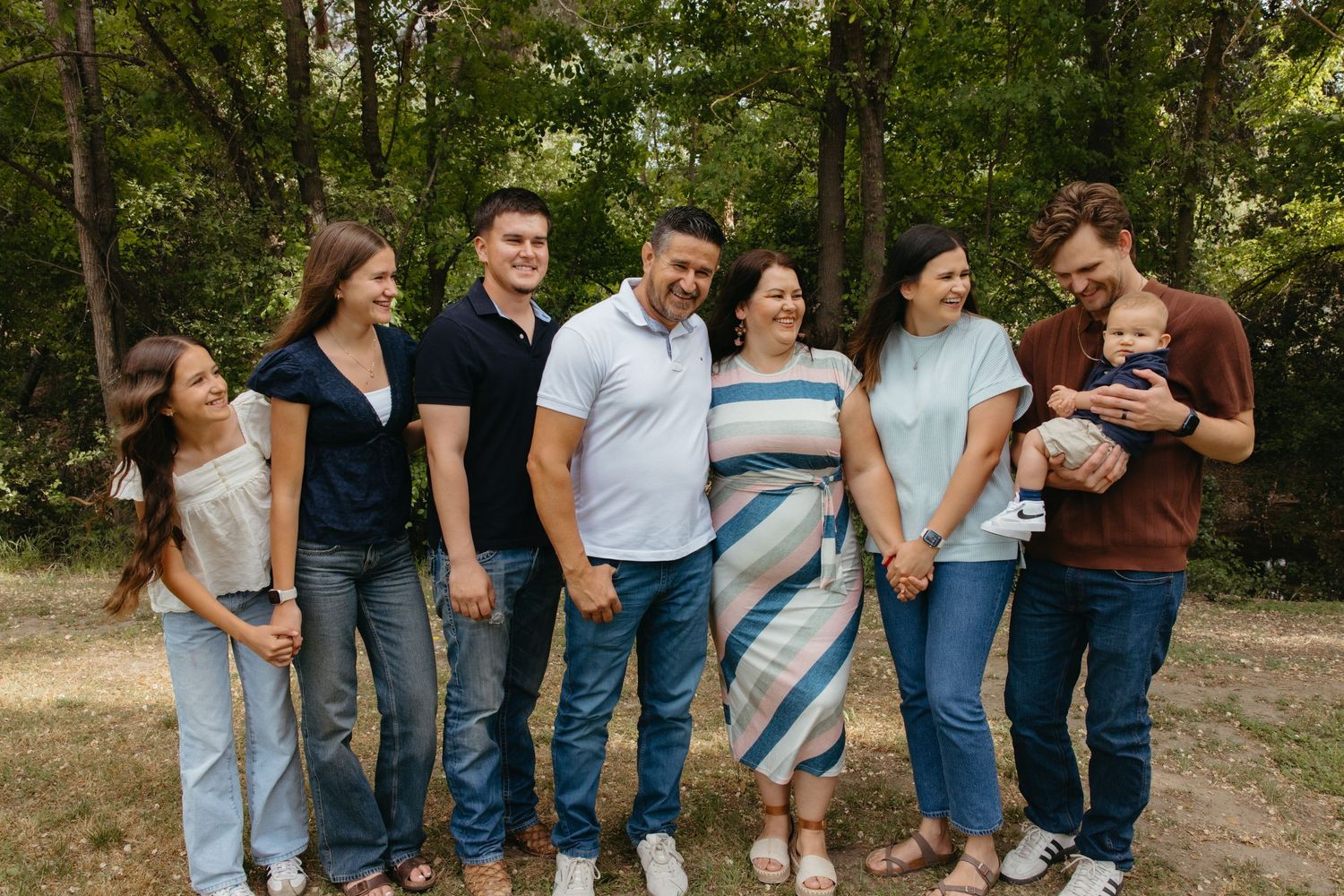 A joyful multigenerational family of seven poses together outdoors surrounded by lush green trees.