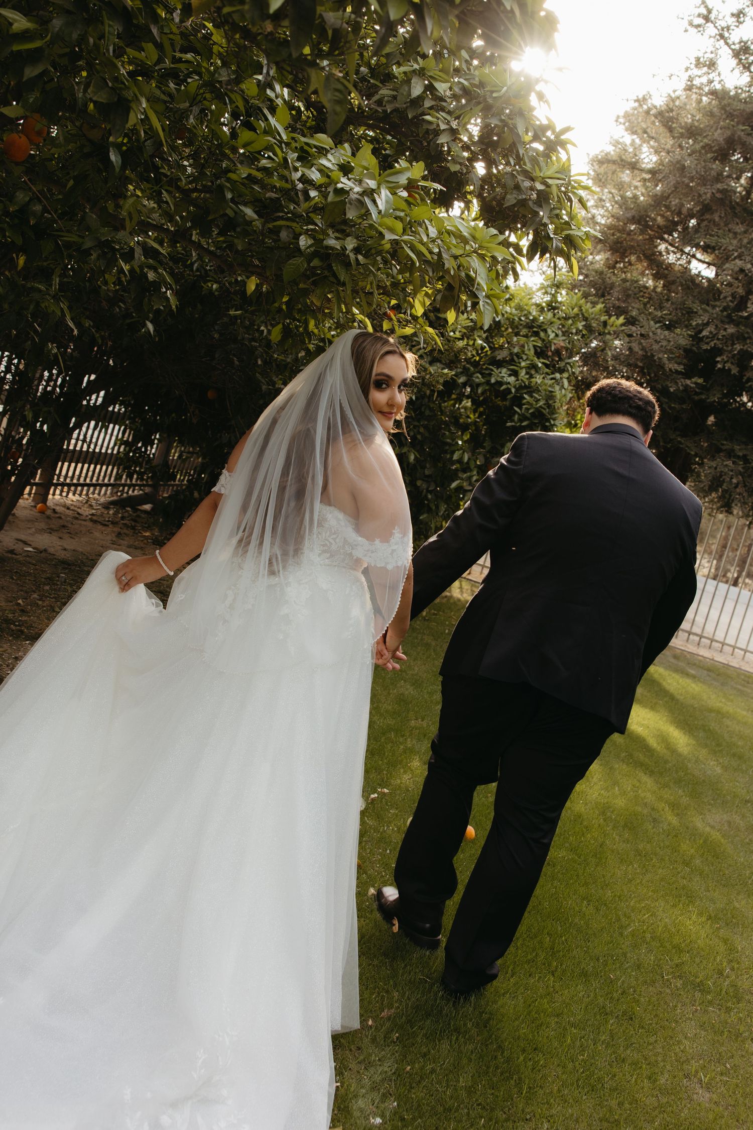 Bride in white ball gown looks back while holding groom's hand walking through a sunlit garden.