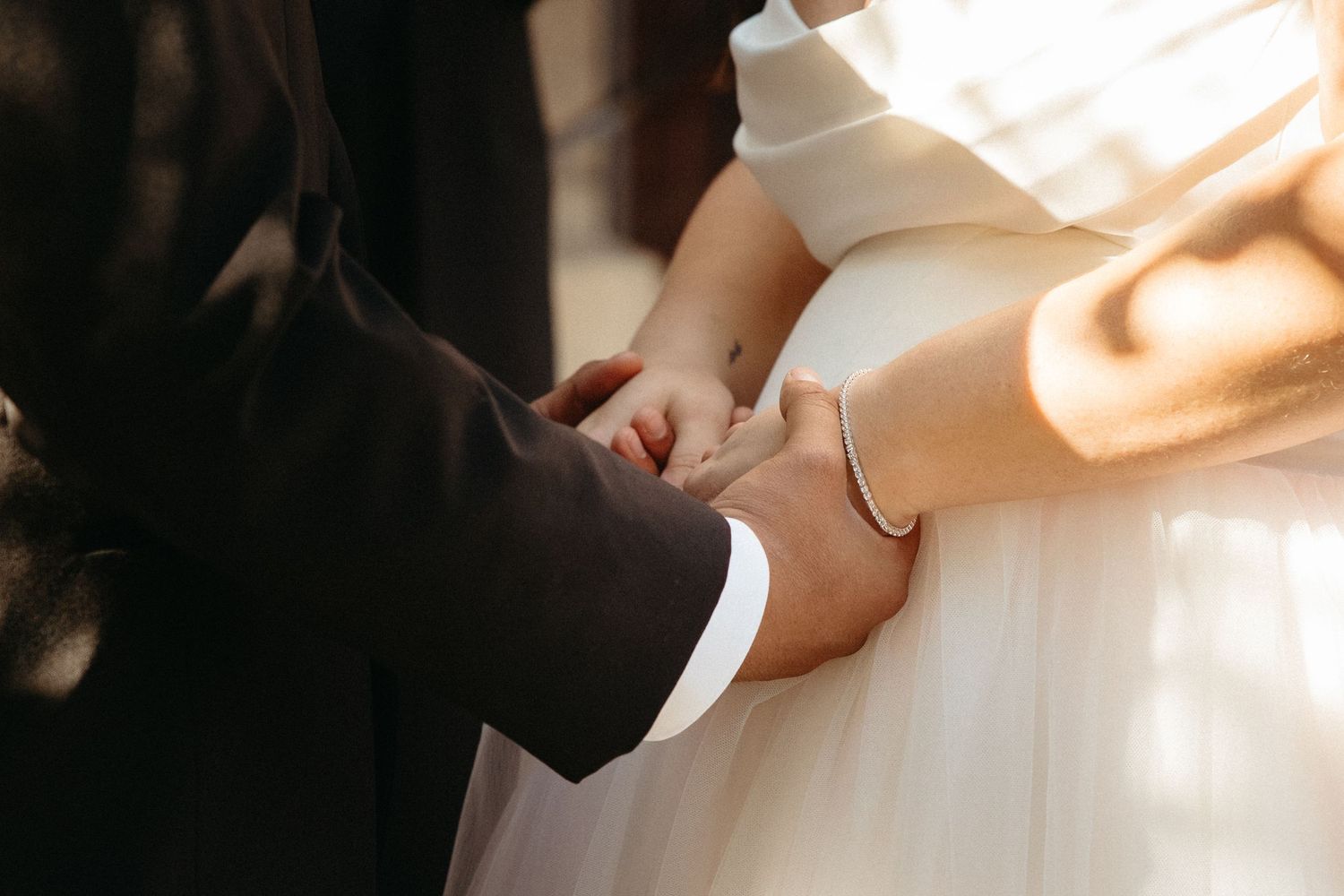 Close-up of a bride and groom holding hands during a sunny wedding ceremony.
