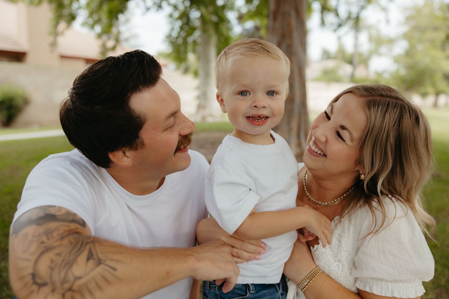 Smiling family of three with tattooed dad, blonde mom, and happy toddler pose outdoors in a park.