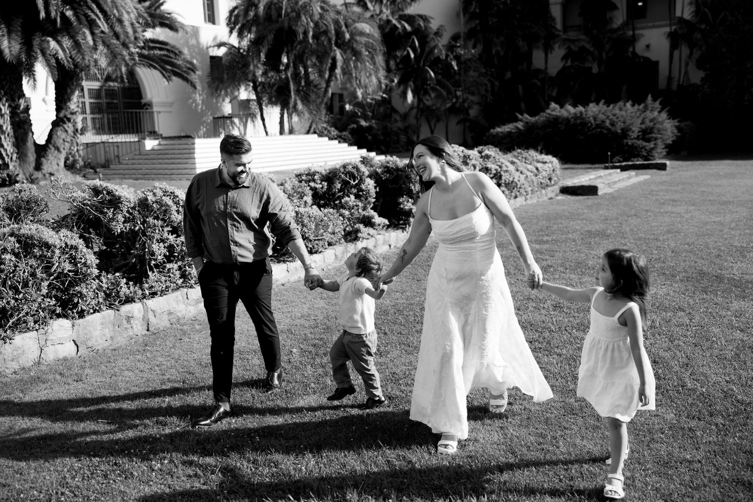 Black and white photo of family of four holding hands and walking together on green lawn.