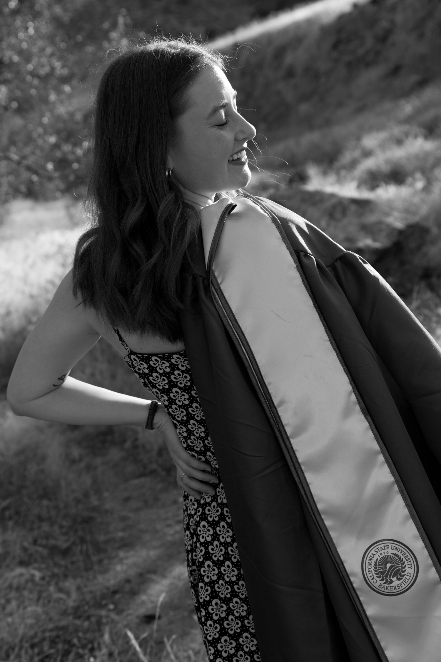 Black and white photo of smiling woman in floral dress holding graduation gown in sunny outdoor setting.