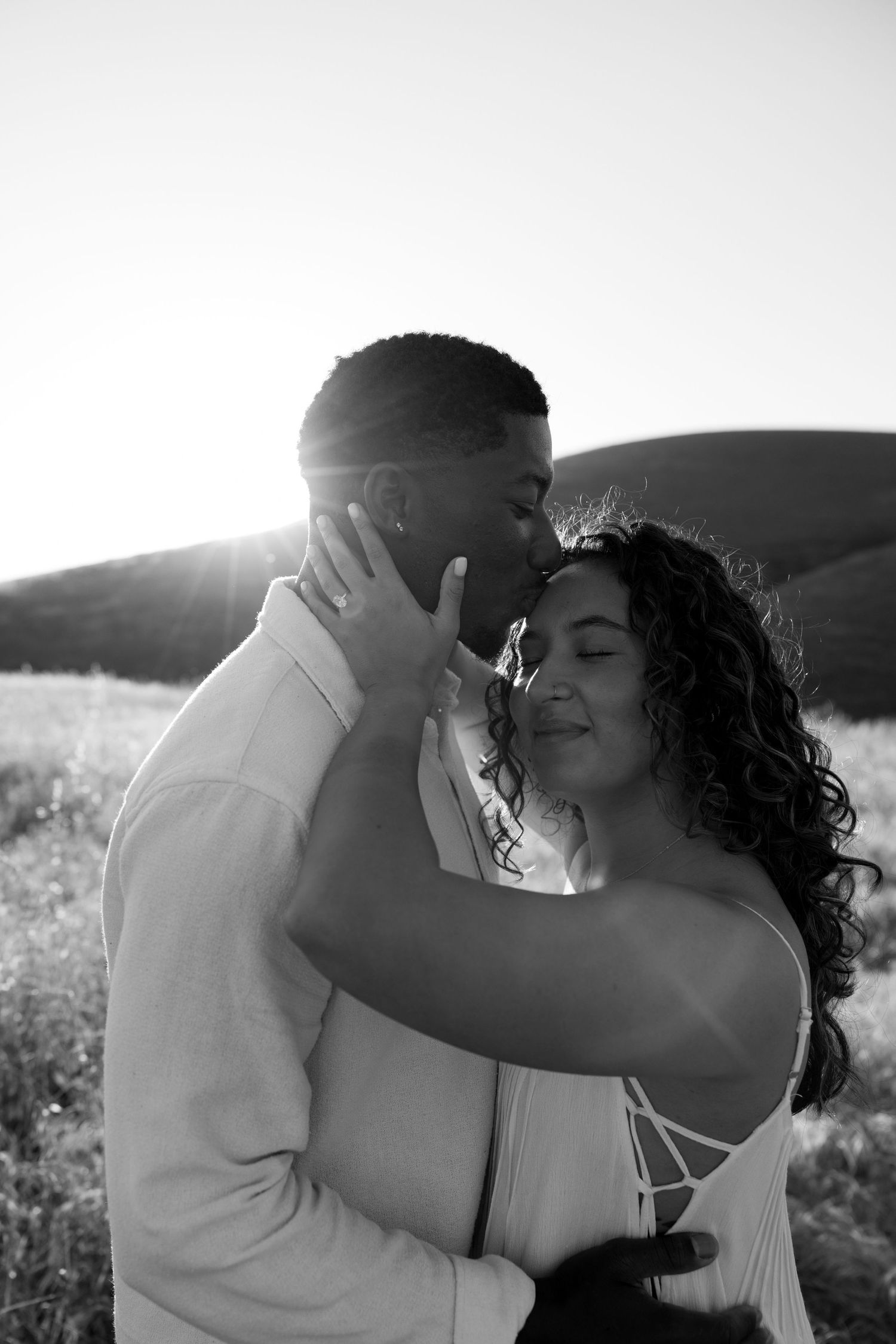 Black and white backlit photo of a couple sharing a tender kiss in an open field at sunset.