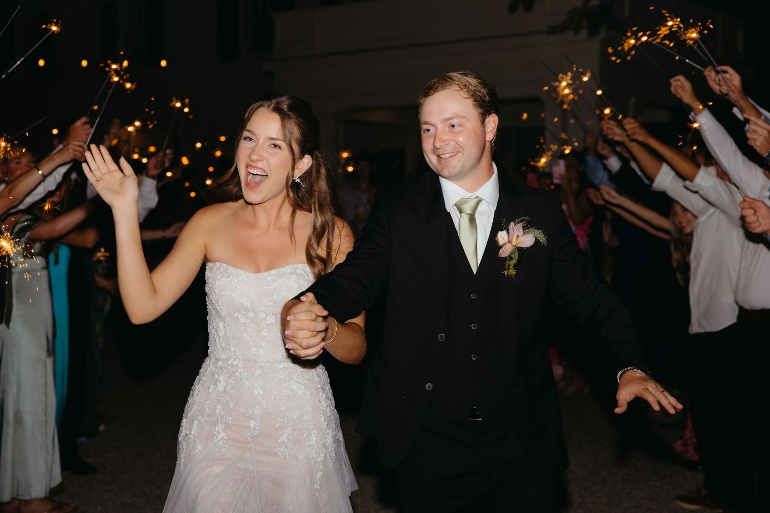 Smiling bride and groom celebrate sparkler exit at night wedding reception with guests cheering.