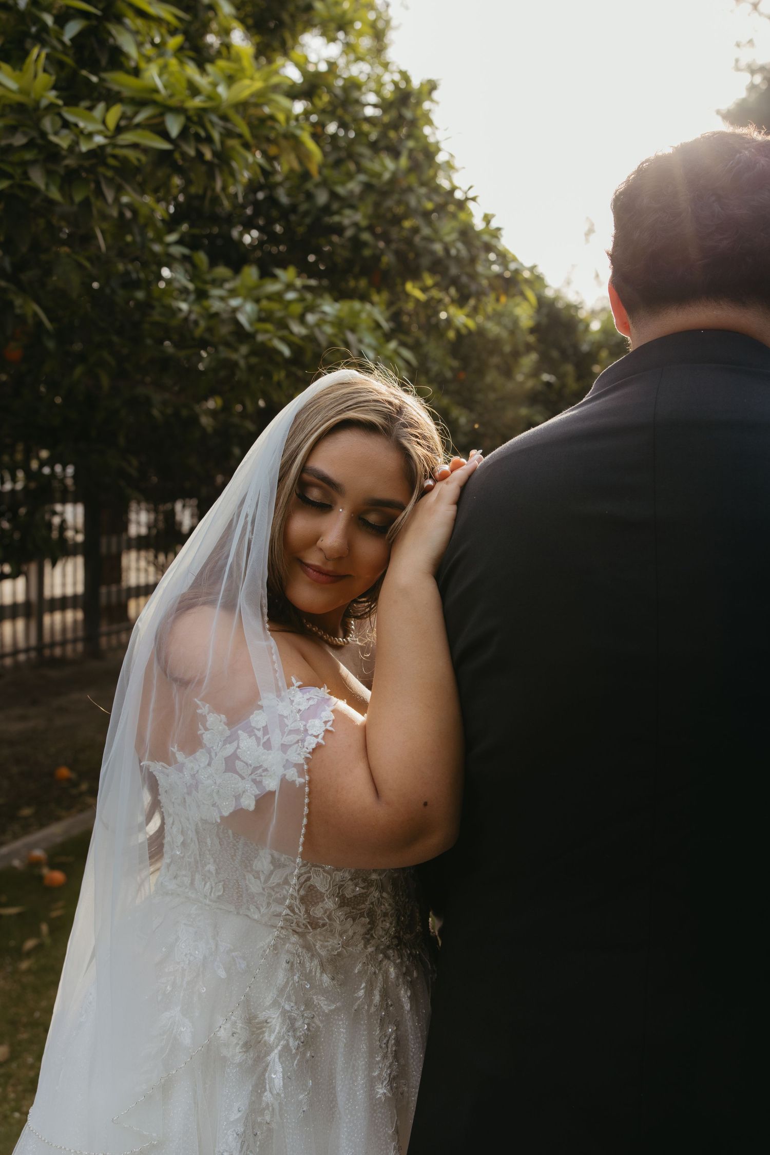 Bride in lace off-shoulder gown rests her head on groom's shoulder in golden sunset light.