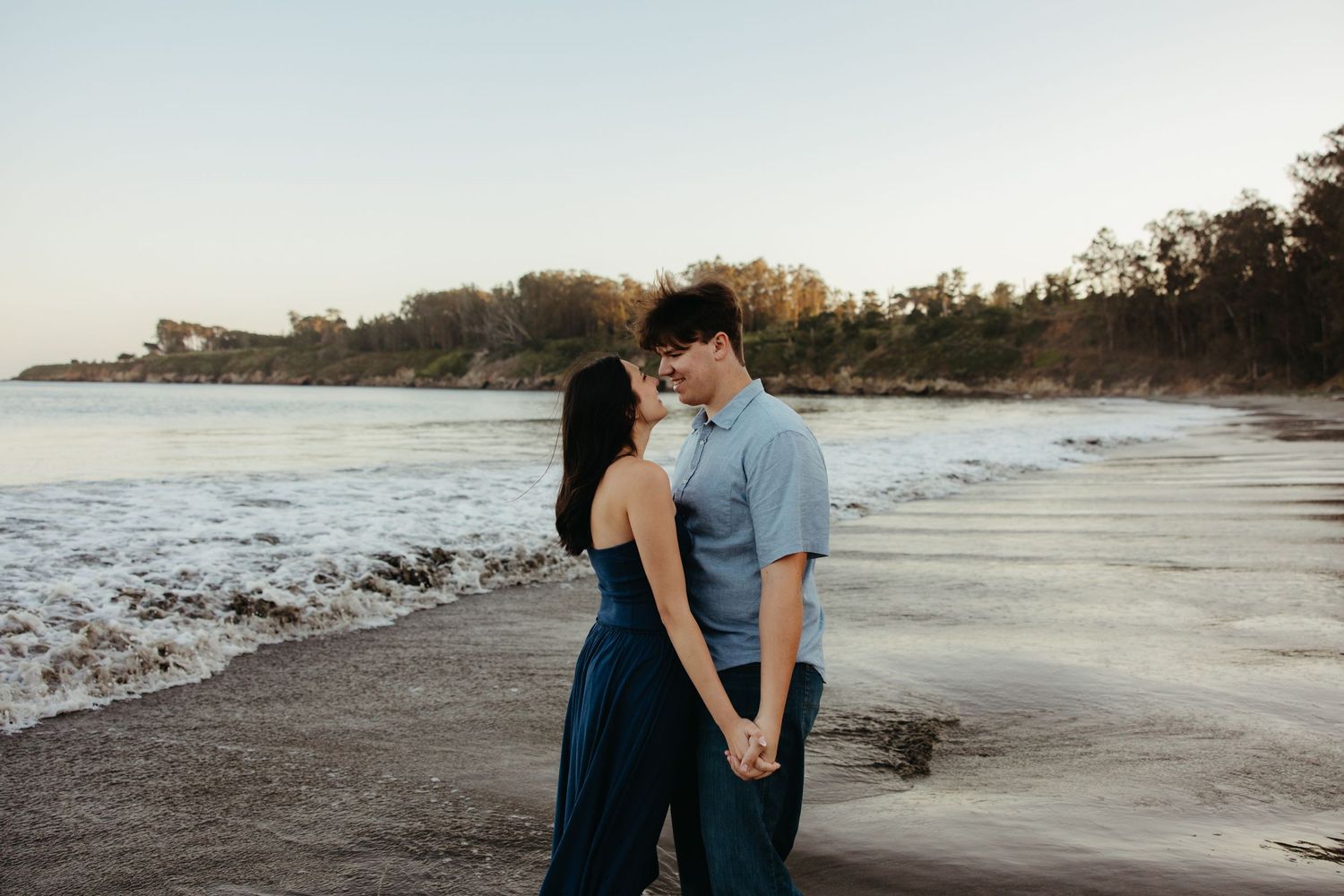 Couple holding hands and sharing a romantic moment on a sandy beach at sunset with ocean waves behind them.