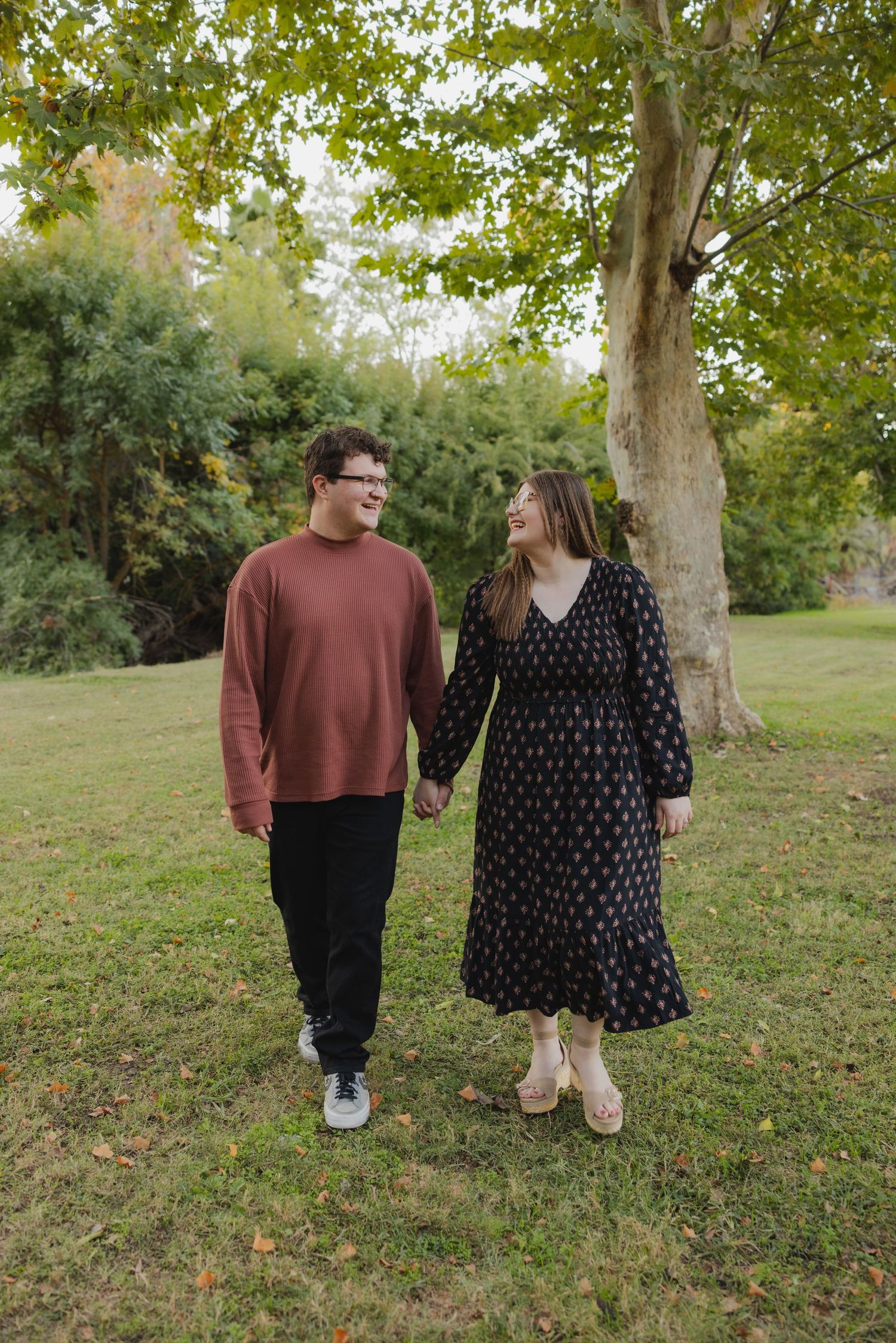 Couple holding hands walking in a green park, man in rust sweater and woman in black floral dress.