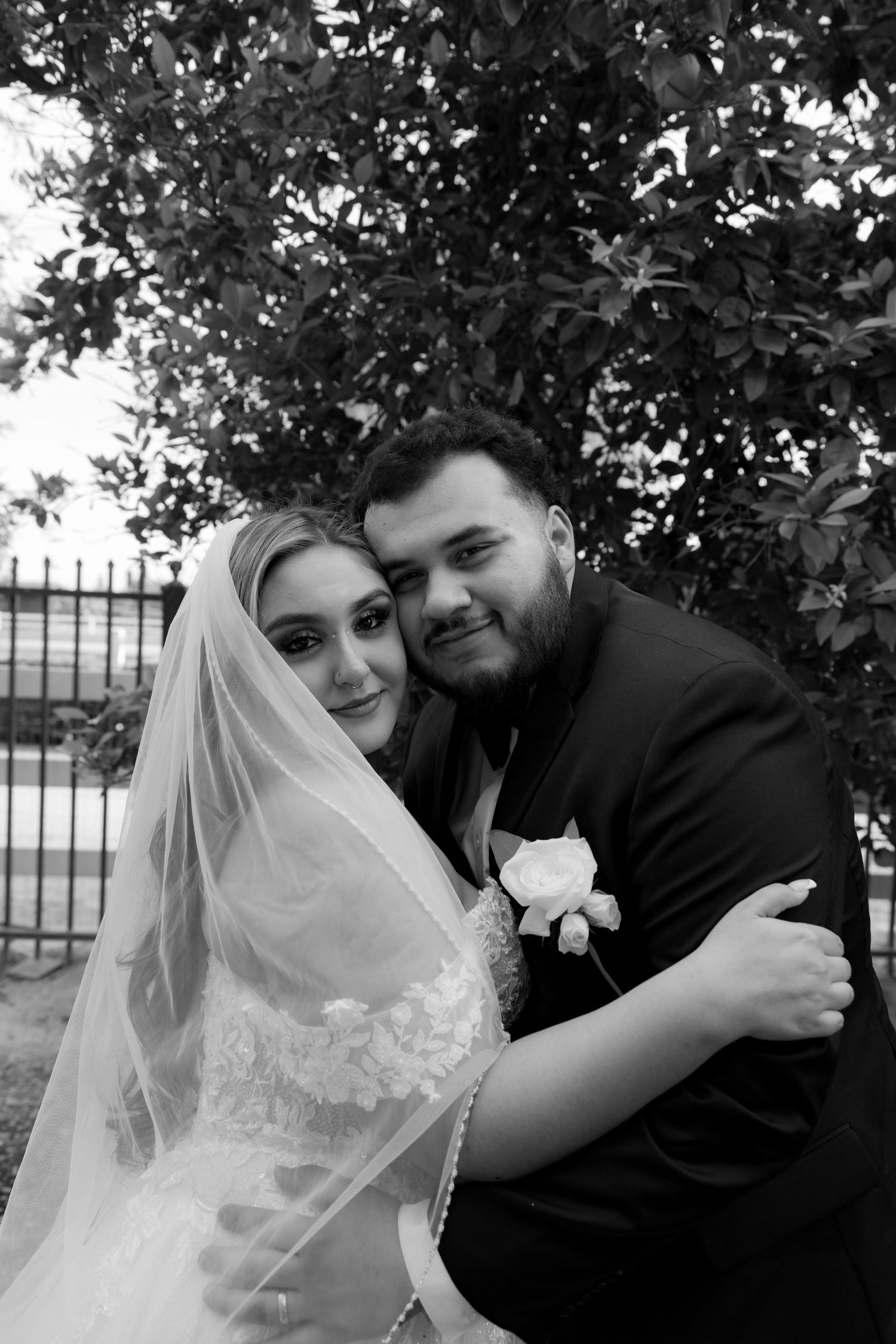 Black and white portrait of smiling bride and groom embracing near a tree after their wedding.