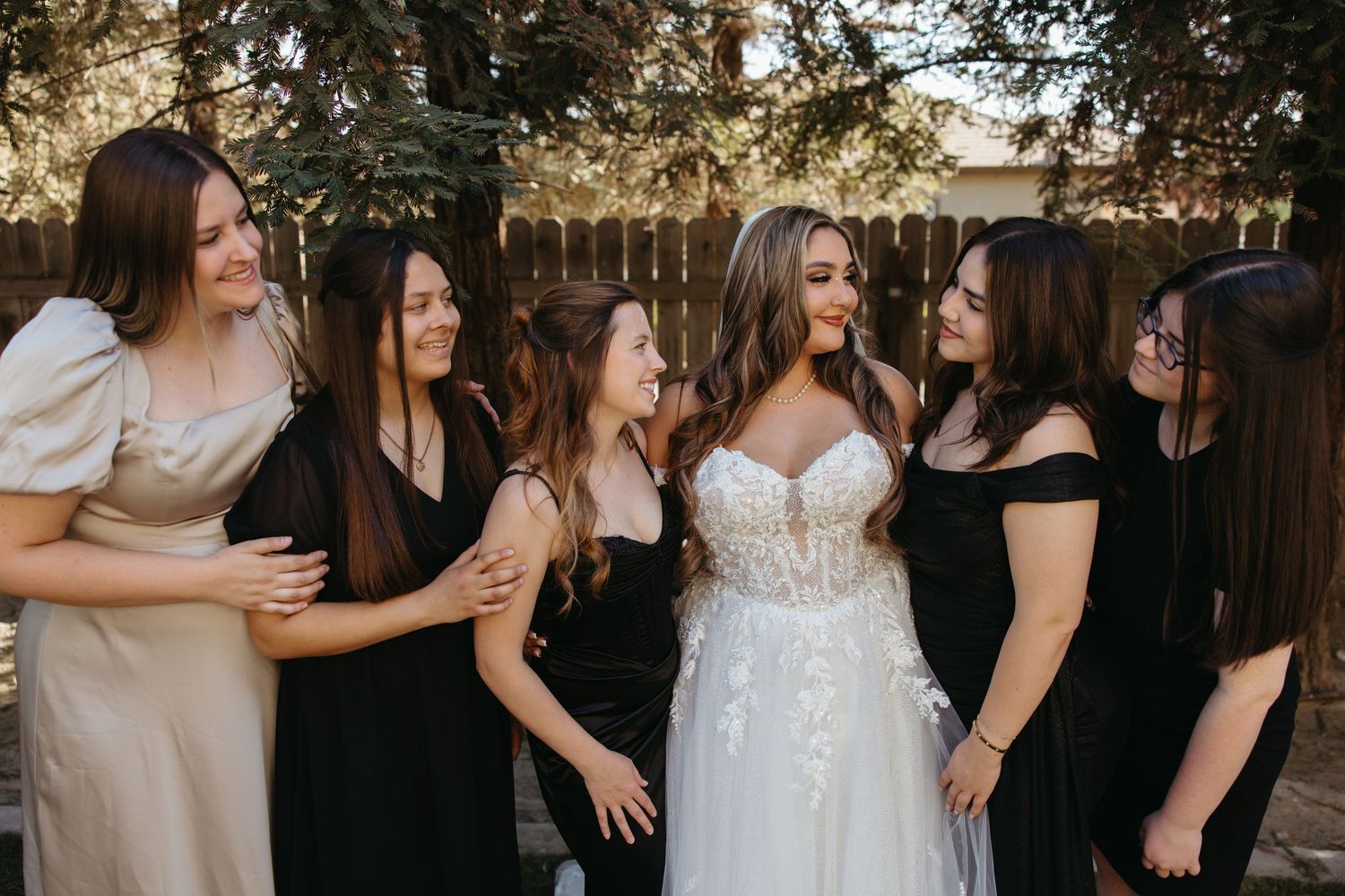 Bride in lace wedding gown laughs with five bridesmaids in black dresses outdoors.