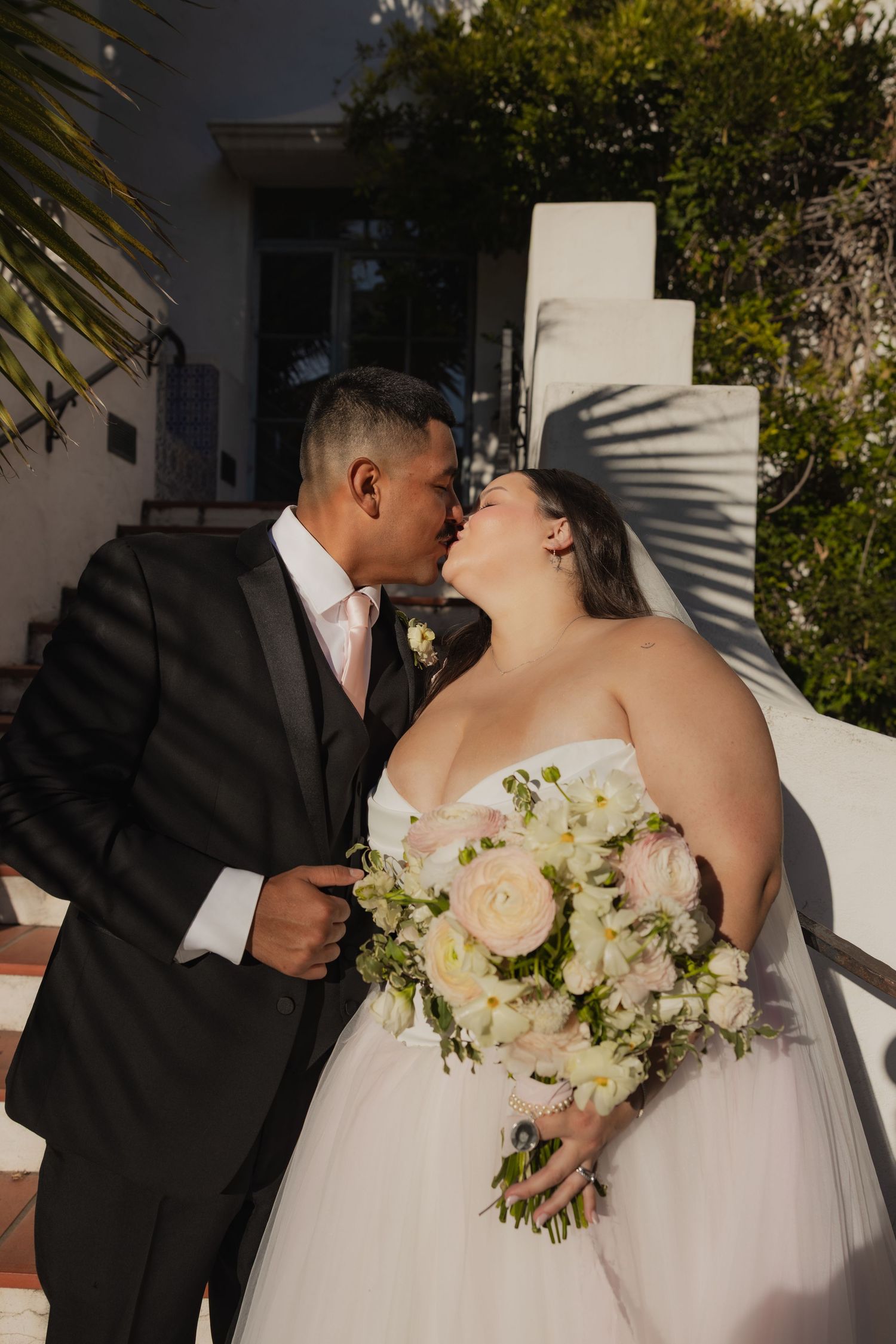 Bride and groom sharing a kiss outdoors, bride holding a bouquet of white and pink roses.