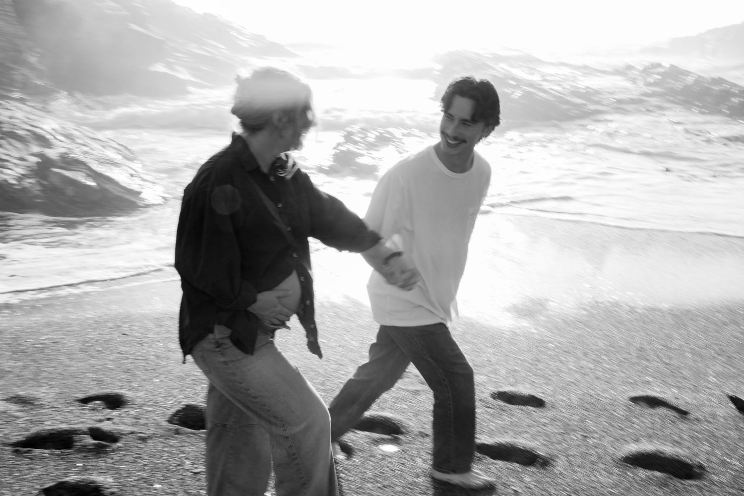 Black and white photo of a couple holding hands and dancing playfully on a misty beach.