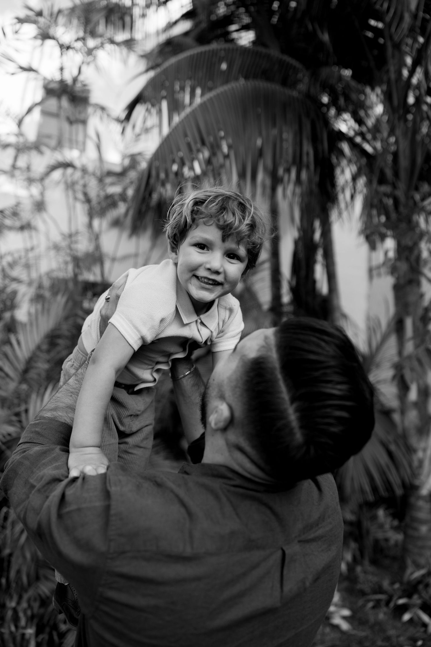 Black and white photo of father lifting laughing toddler boy near palm trees outdoors.